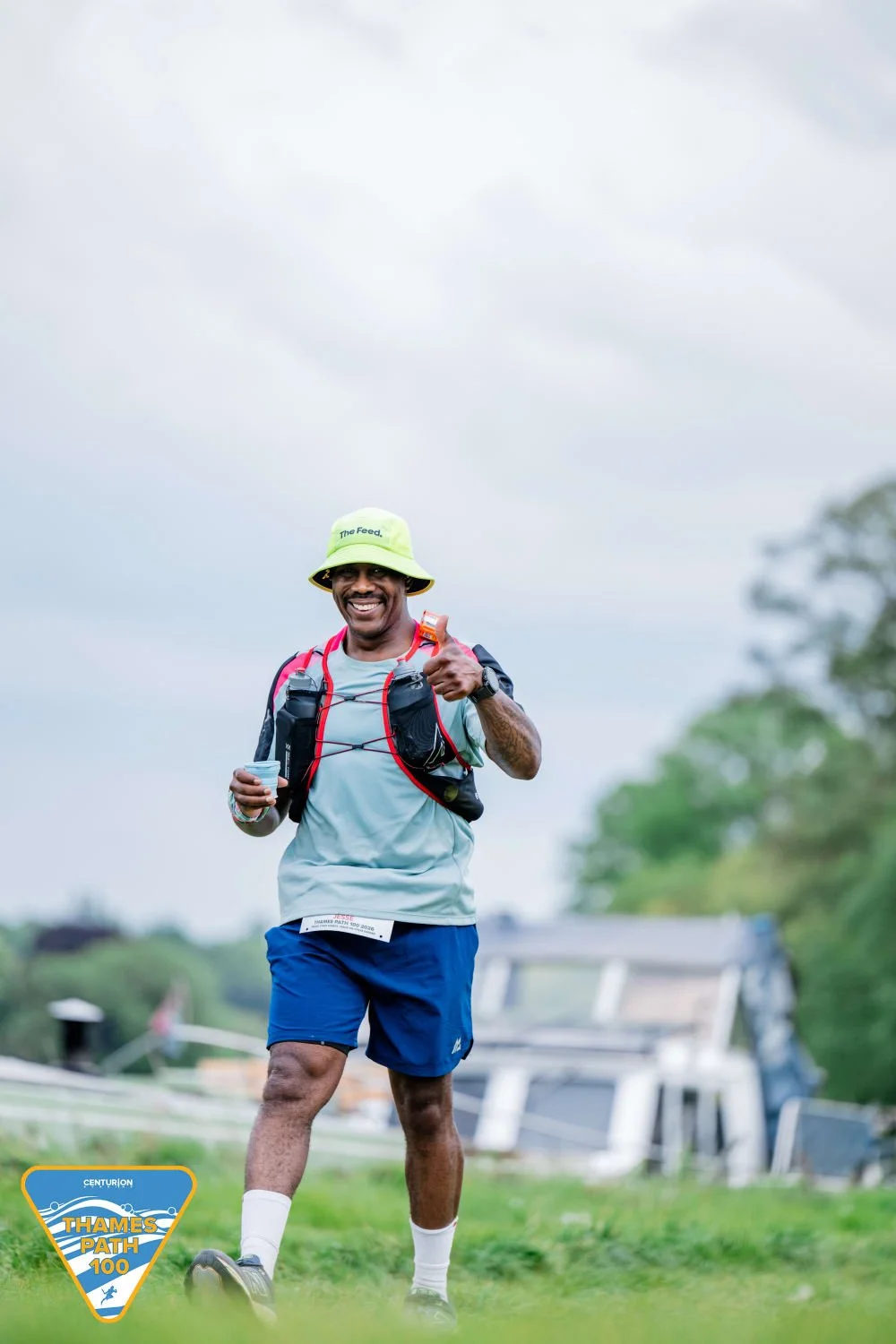 A smiling man in running gear and a yellow hat giving a thumbs-up during a race outdoors.