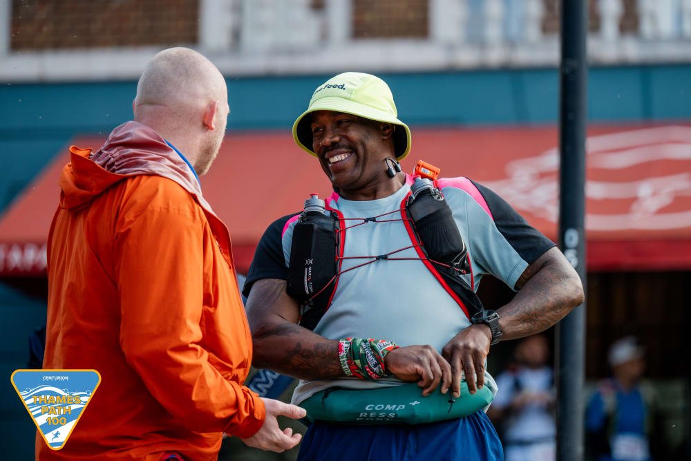 Two men talking outdoors, one wearing an orange jacket and the other wearing a gray t-shirt, a yellow hat, and a hydration pack, smiling and engaging in conversation.