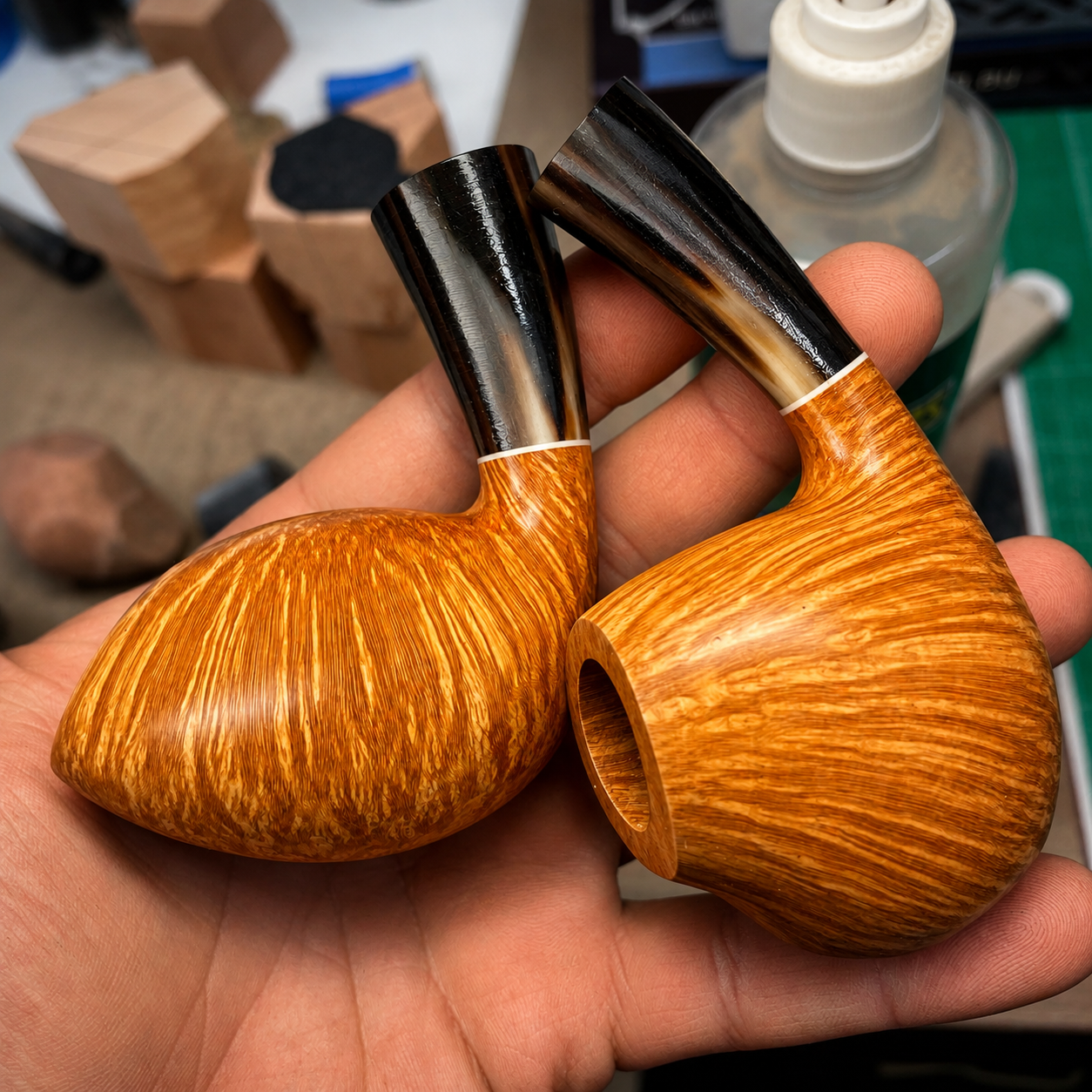 A close-up of two finely crafted wooden smoking pipes with a glossy finish, showing detailed wood grain patterns. The pipes feature curved designs with black and brown mouthpieces and are held in a person's hand. The background includes blurred woodw
