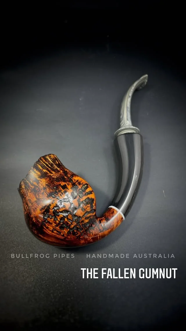 A handcrafted smoking pipe with a decorated bowl and a curved gray stem on a dark background, advertising Bullfrog Pipes from Australia, called The Fallen Gunnut.
