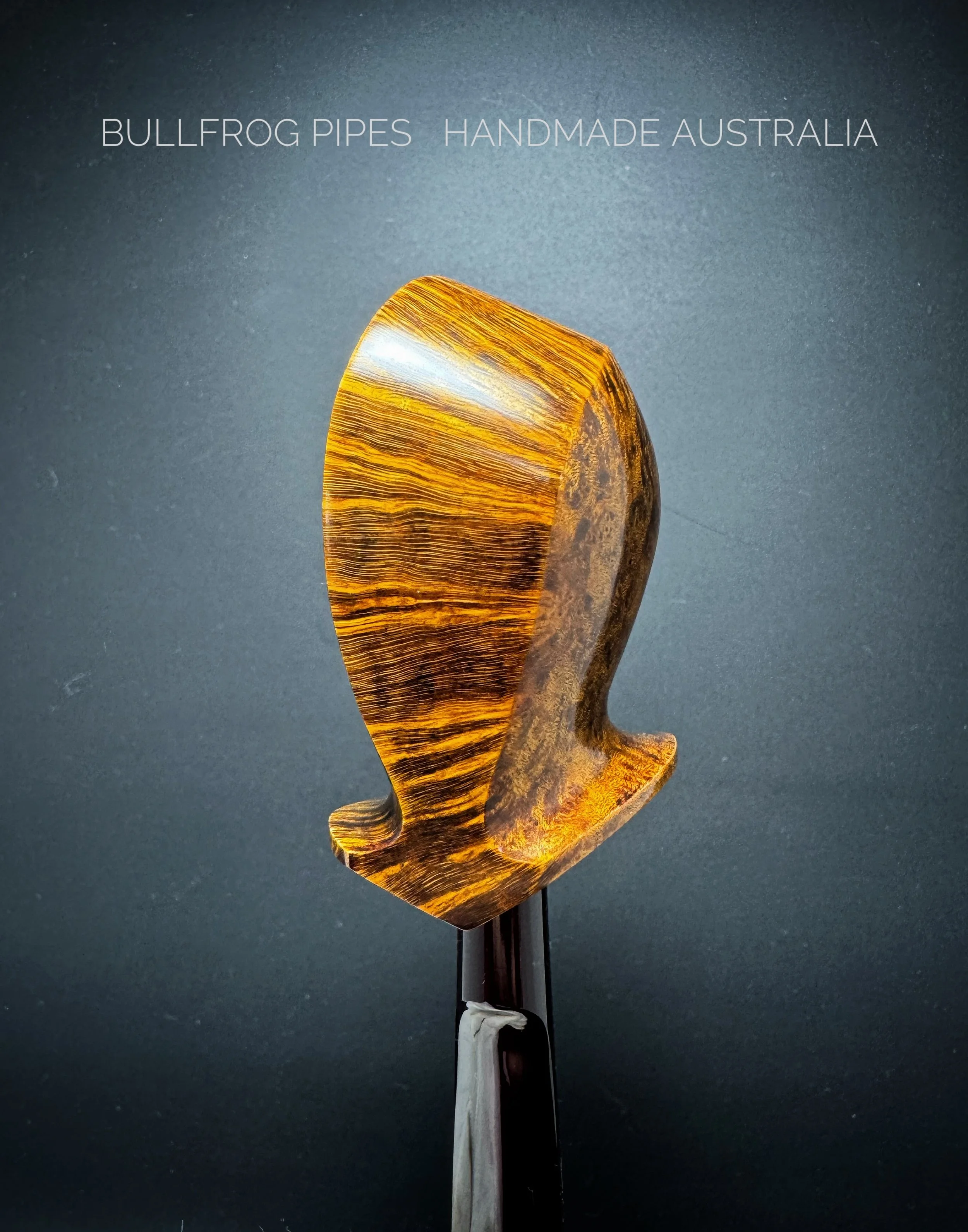 A wooden sculpture of a bullfrog pipe with a yellow and brown striped pattern, mounted on a black stand against a dark background.