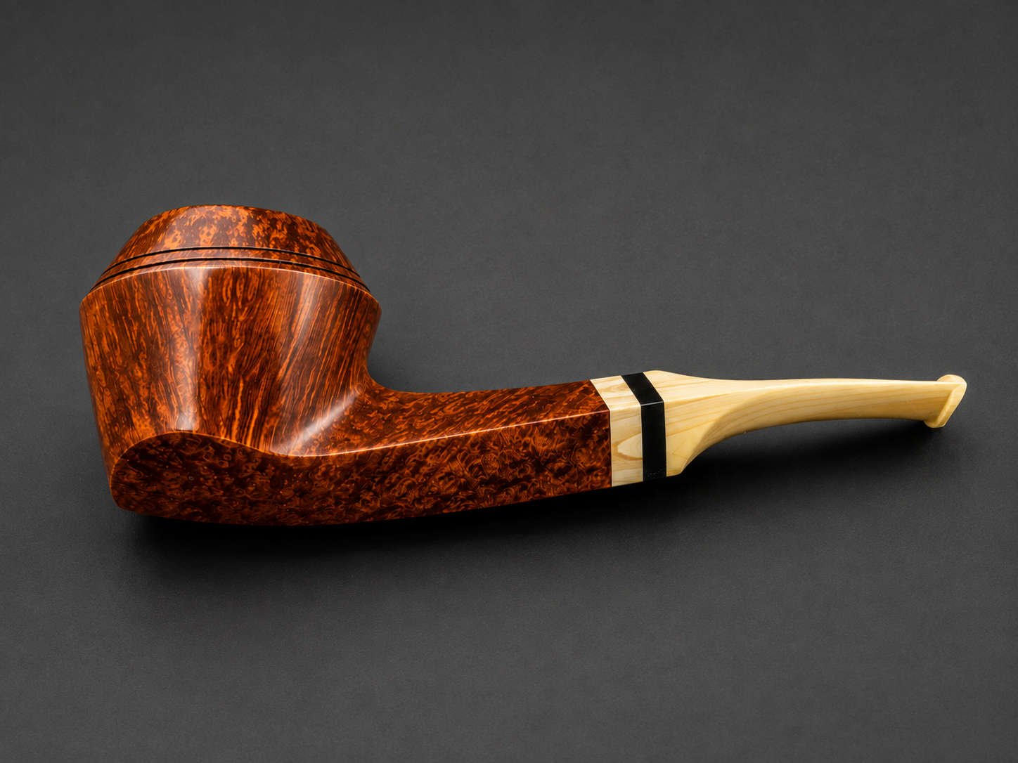 A close-up view of a wooden tobacco pipe with a curved stem, featuring a two-tone design with dark, intricate grain on the bowl and a light-colored stem separated by a black band, against a dark background.