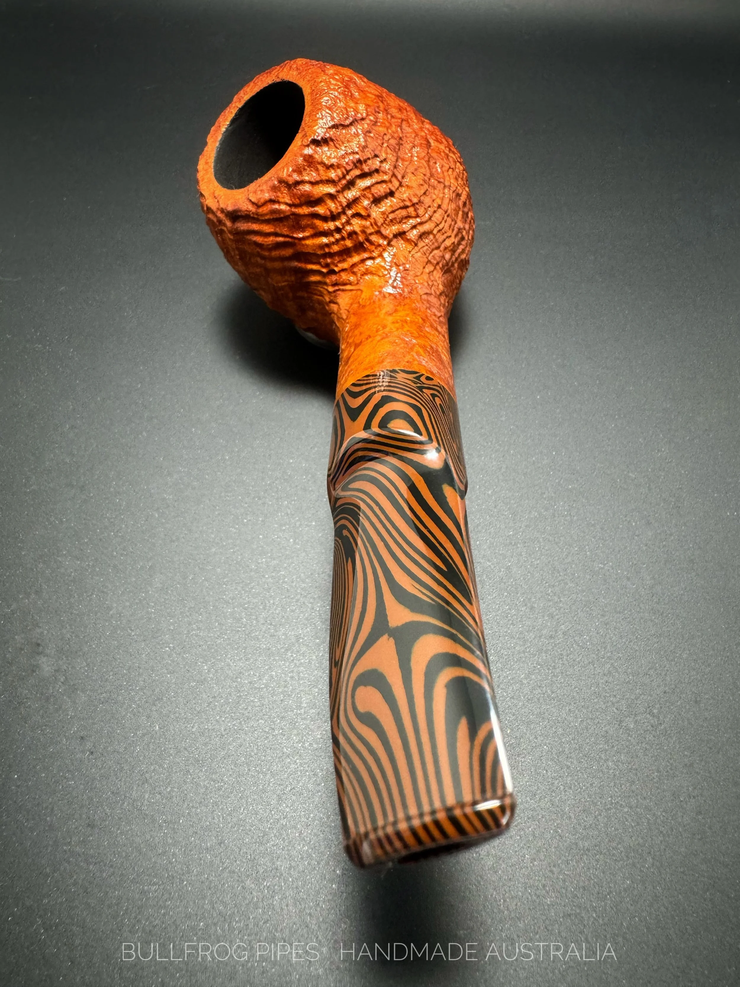 A handmade Australian pipe with a textured orange and black design on the stem, and a rough, cracked orange bowl. The pipe is resting on a dark surface, and text at the bottom reads 'BULLFROG PIPES HANDMADE AUSTRALIA'.