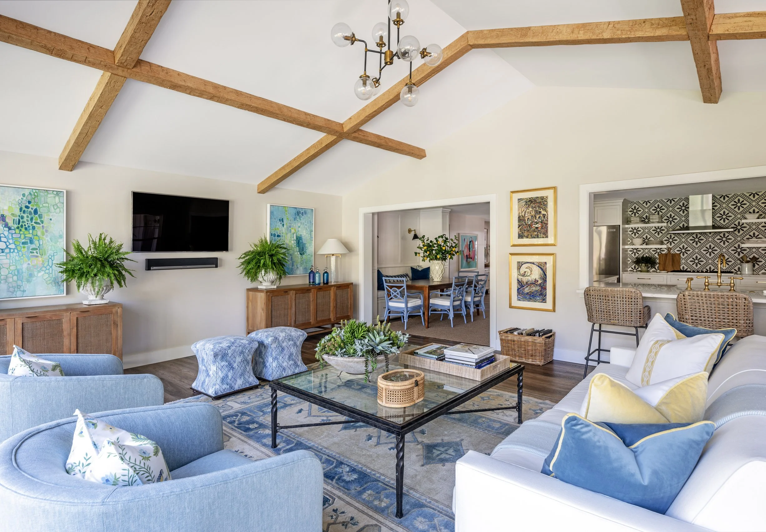 A living room with light-colored walls, wooden beamed ceiling, a flat-screen TV, potted plants, a modern chandelier, colorful paintings, and a seating area with blue and white upholstered furniture.