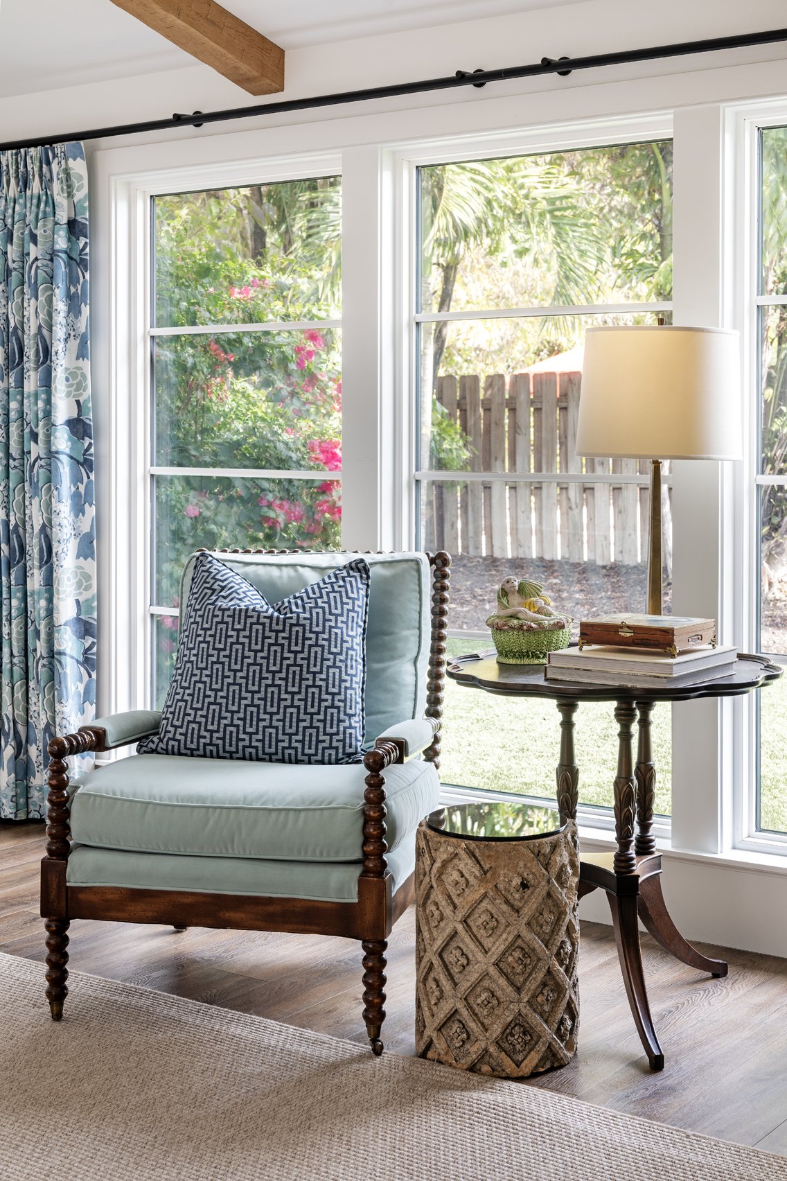 Living room with large windows, a light blue armchair with a patterned pillow, a small wooden side table with decorative items, a table lamp, and a view of greenery outside.
