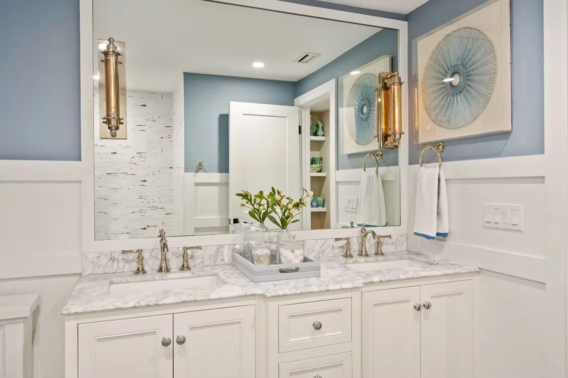 A bathroom vanity with a marble countertop, a large mirror, two wall-mounted gold light fixtures, and decorative artwork on the wall. There are two white towels hanging on gold towel rings and a tray with toiletries on the countertop.