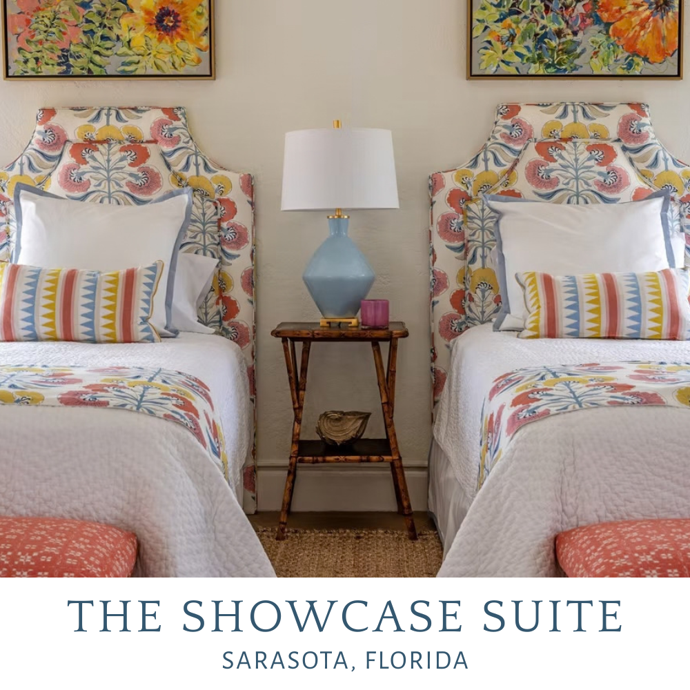 Two twin beds with colorful floral and geometric patterned headboards, white bedding, and matching decorative pillows, a wooden nightstand with a blue lamp, a pink candle, and a purple container between the beds, with framed floral artwork above.