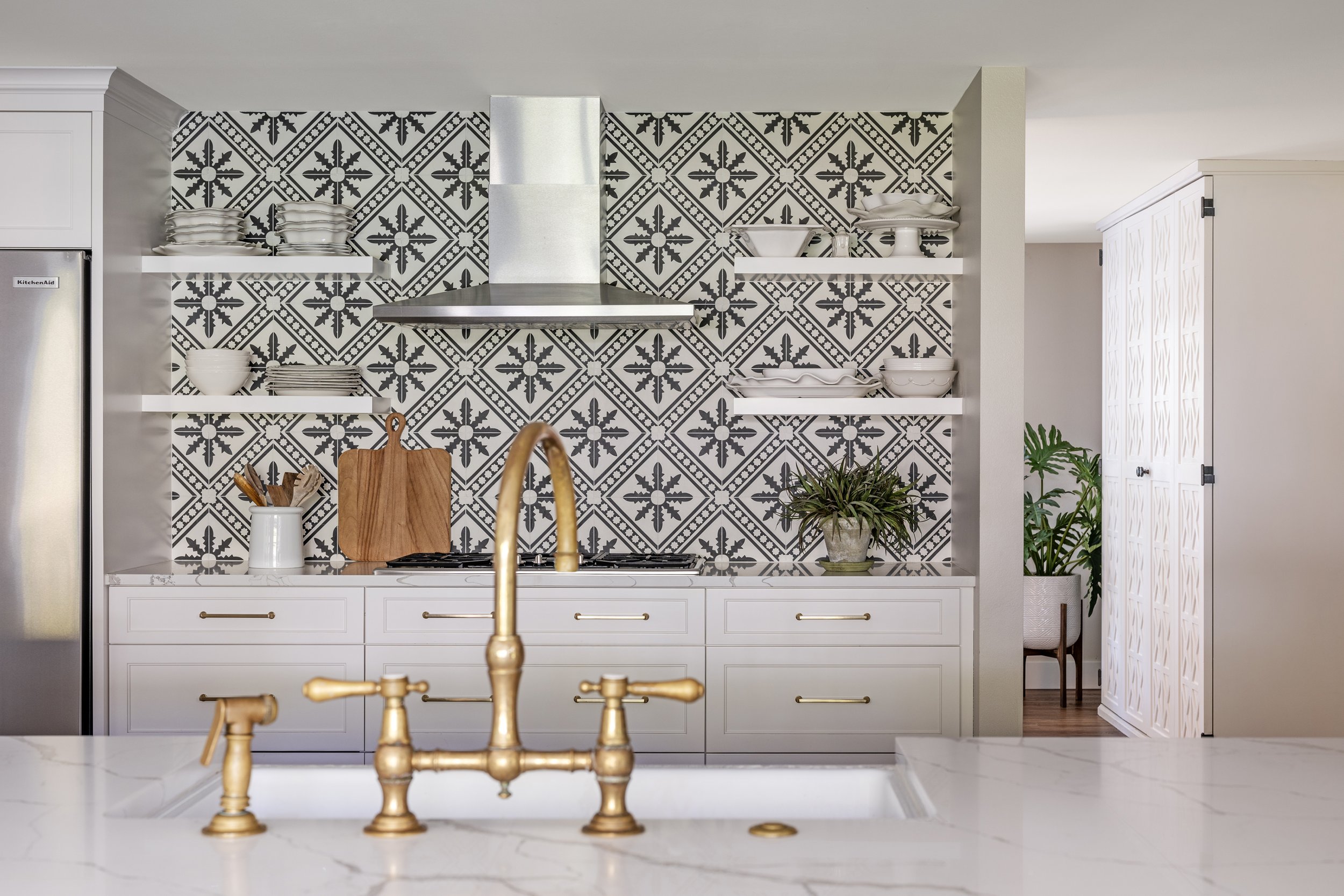 Kitchen with black and white patterned tile backsplash, white cabinets, open shelves with dishes, brass faucet, and potted plants.