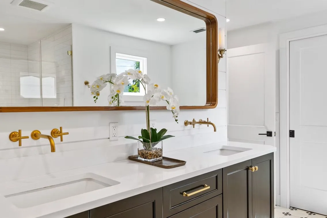 A modern bathroom vanity with a large mirror, black cabinets with gold hardware, white countertop, two sinks, gold fixtures, an orchid plant in a glass container, and a window in the background. Neutral bathroom designed by Dalton Reed Interiors.
