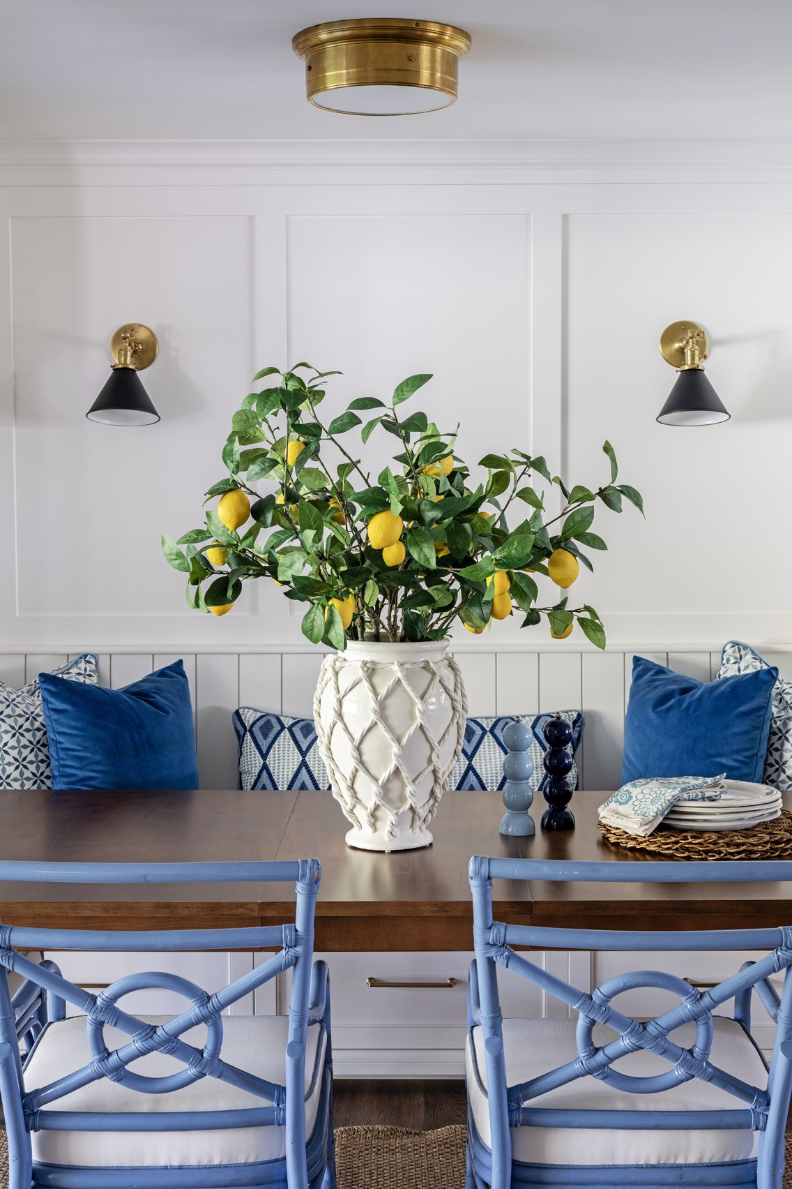 A dining table with blue chairs, a large potted lemon tree with yellow lemons, blue and white pillows on a bench, and wall-mounted black and gold lamps in a bright, modern dining room.