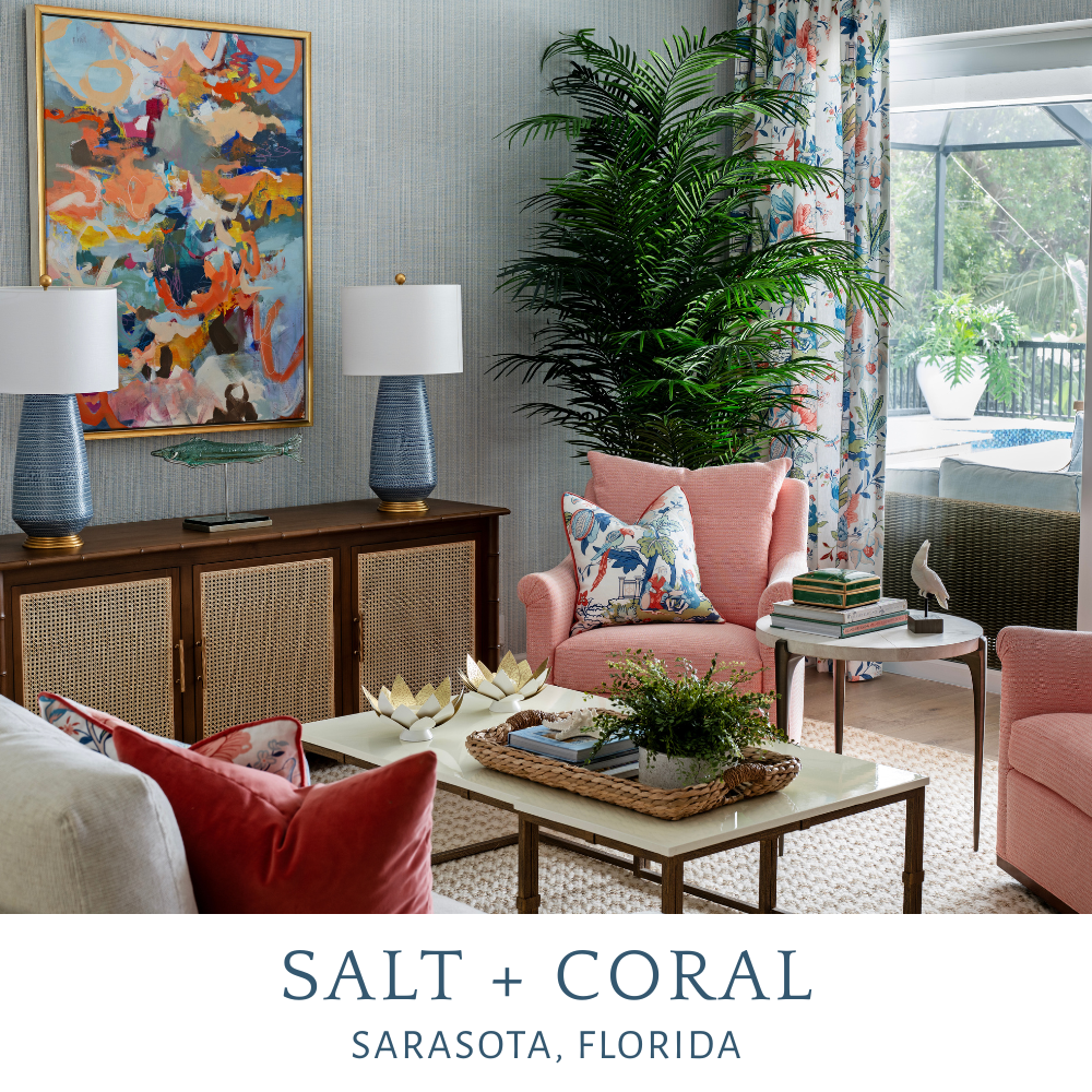 A living room with a pastel color scheme, featuring pink armchairs, a white coffee table with decorative candles, a wicker tray, a plant, and a colorful abstract painting on the wall. There are two lamps on a wooden console, a large green potted plan