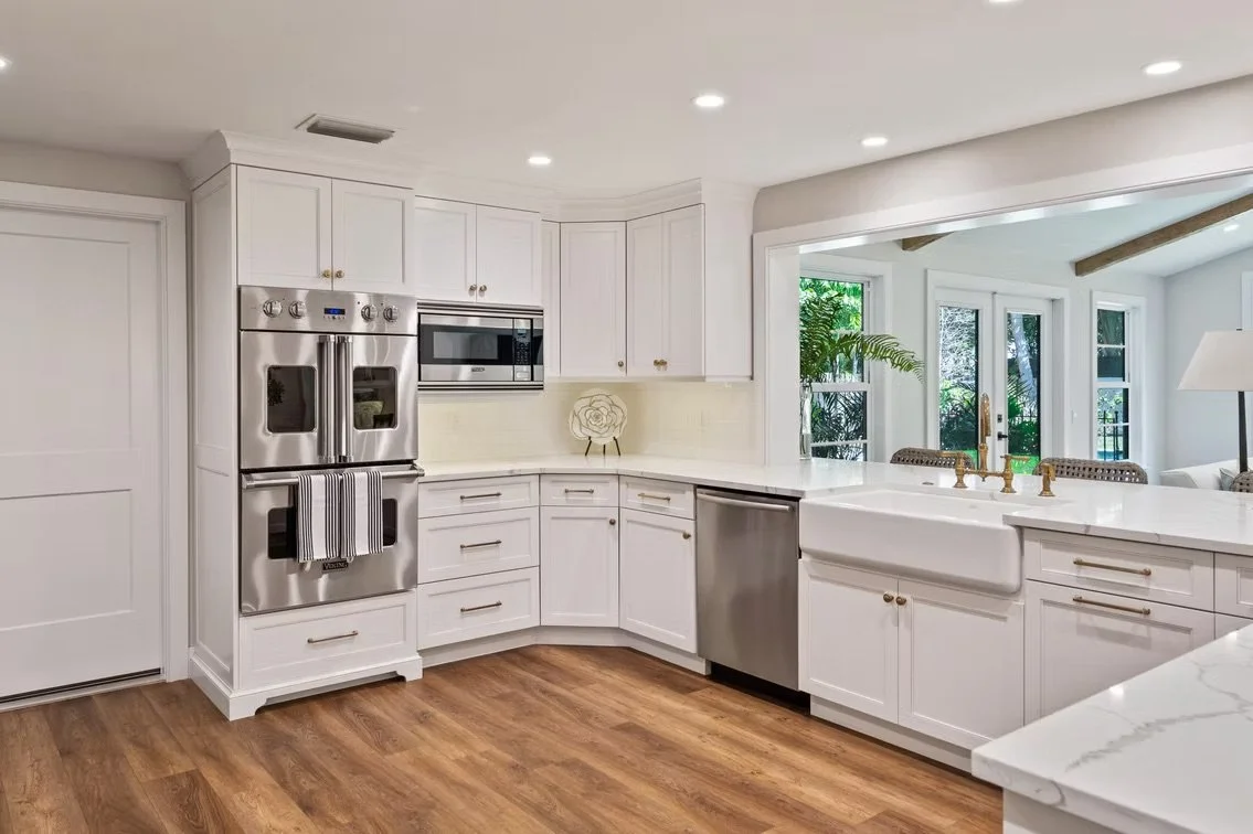 Modern white kitchen with wood flooring, stainless steel appliances, and a view to a sunlit living area with large windows and a sliding door.