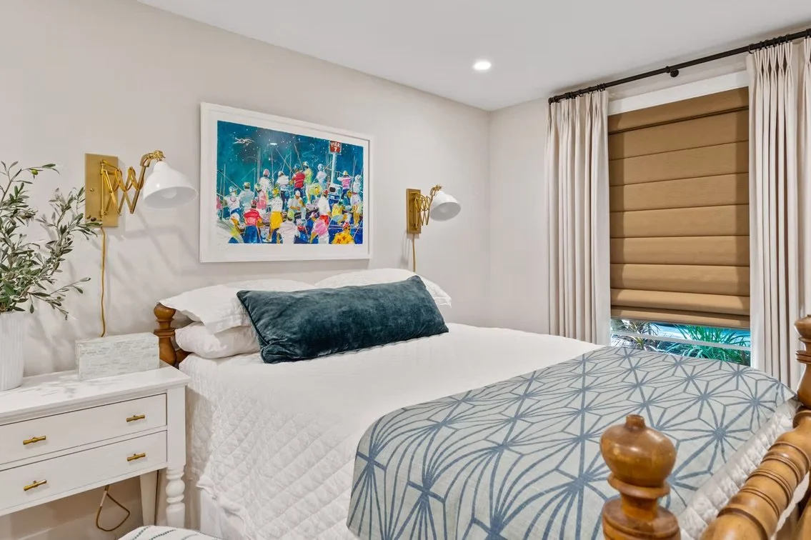 A cozy bedroom with a white bed, dark teal pillow, wall-mounted lamps, framed colorful artwork, beige curtains, and a window with blinds.
