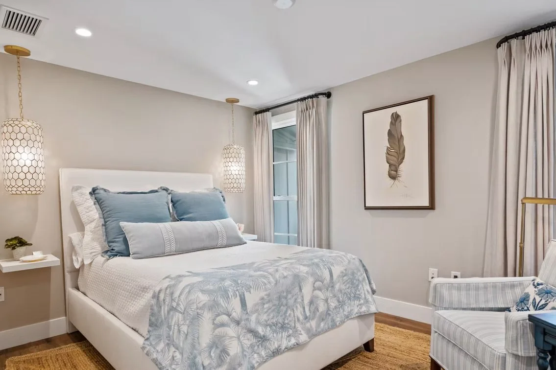 A bedroom with a white bed, blue and white pillows, beige walls, beige curtains, a framed feather print, and a white armchair.