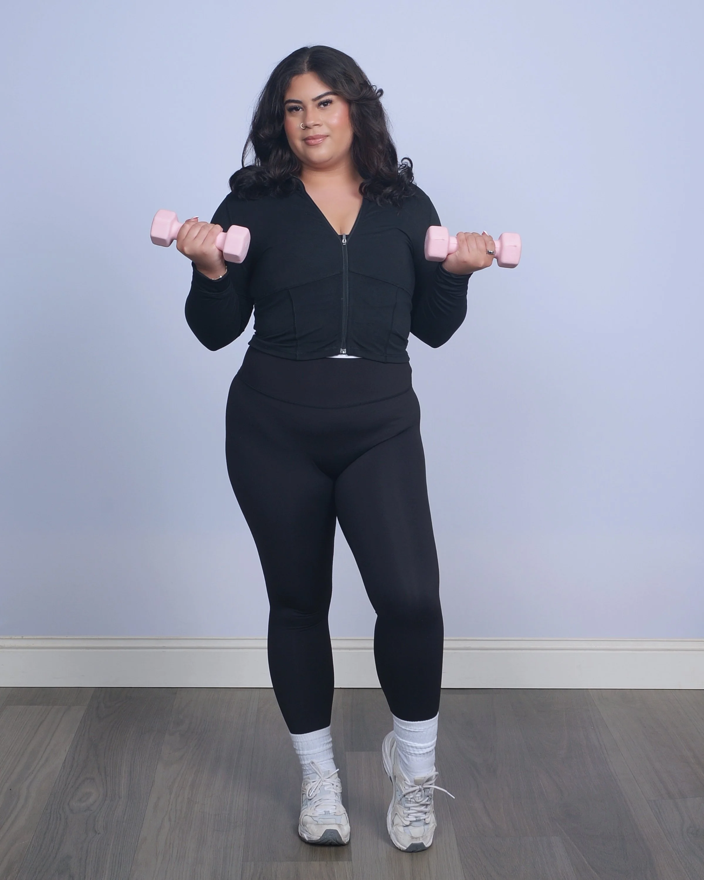 A woman with dark, curly hair holding pink dumbbells in both hands, standing against a light blue wall. She is wearing a black zip-up athletic jacket, black leggings, white socks, and white sneakers.