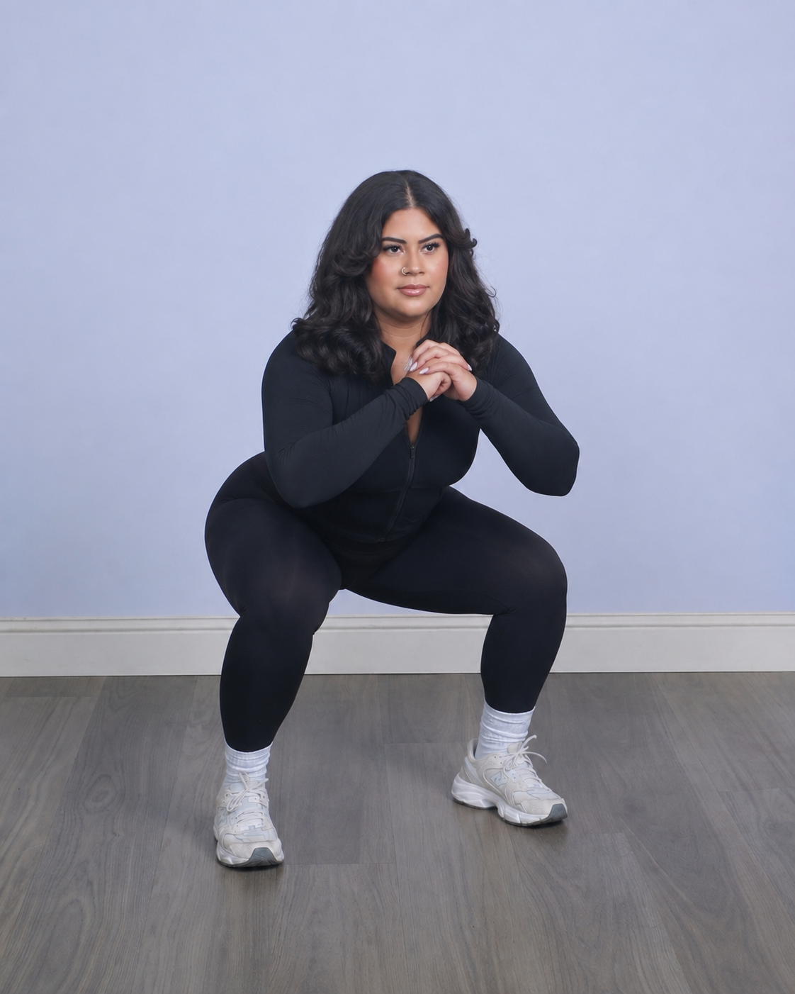 Woman in black athletic wear squatting indoors with hands clasped in front of her, on a wooden floor with plain light-colored wall background.