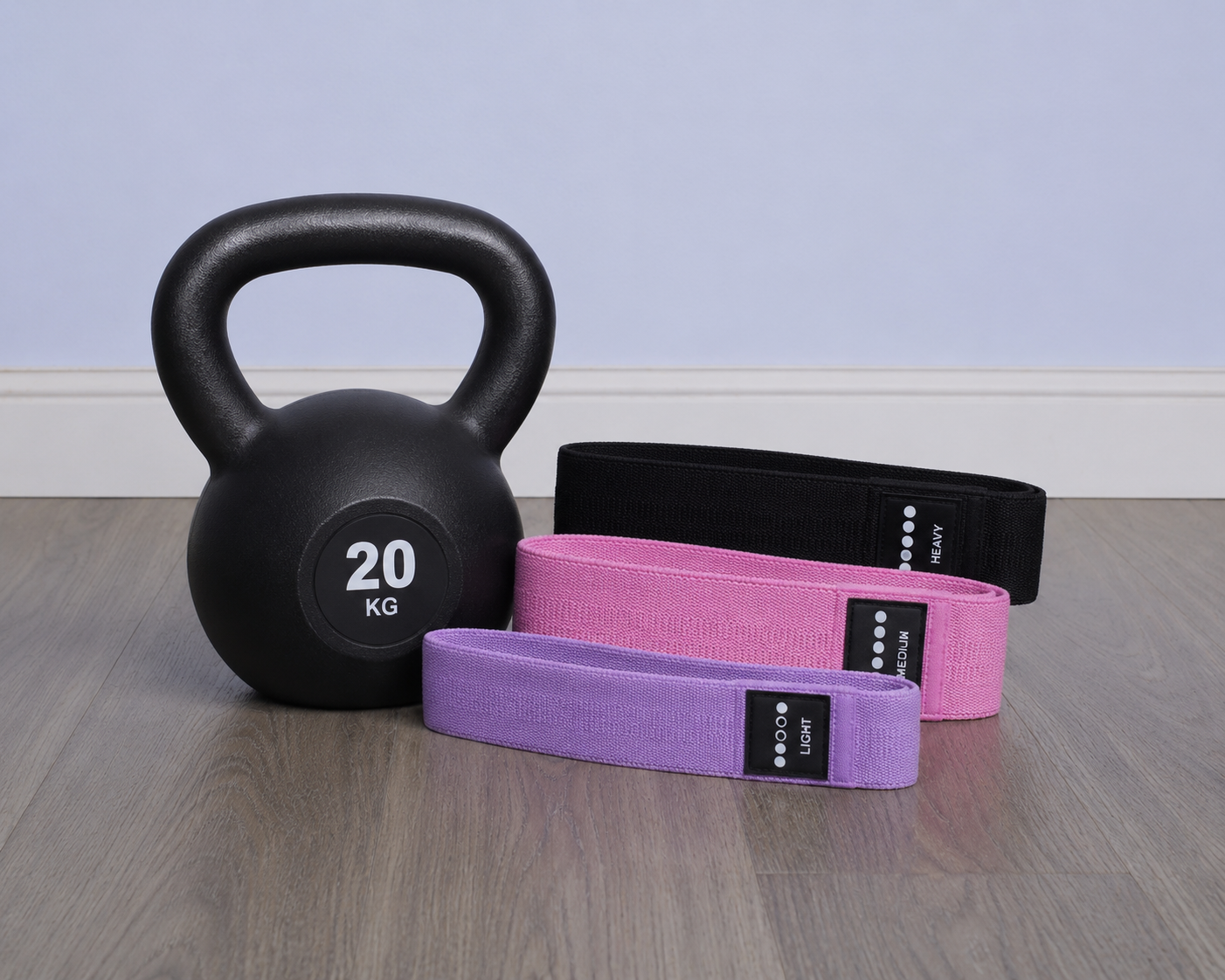 A black kettlebell with a 20 kg label, three resistance bands in black, pink, and purple, all arranged on a wooden floor against a light-colored wall.