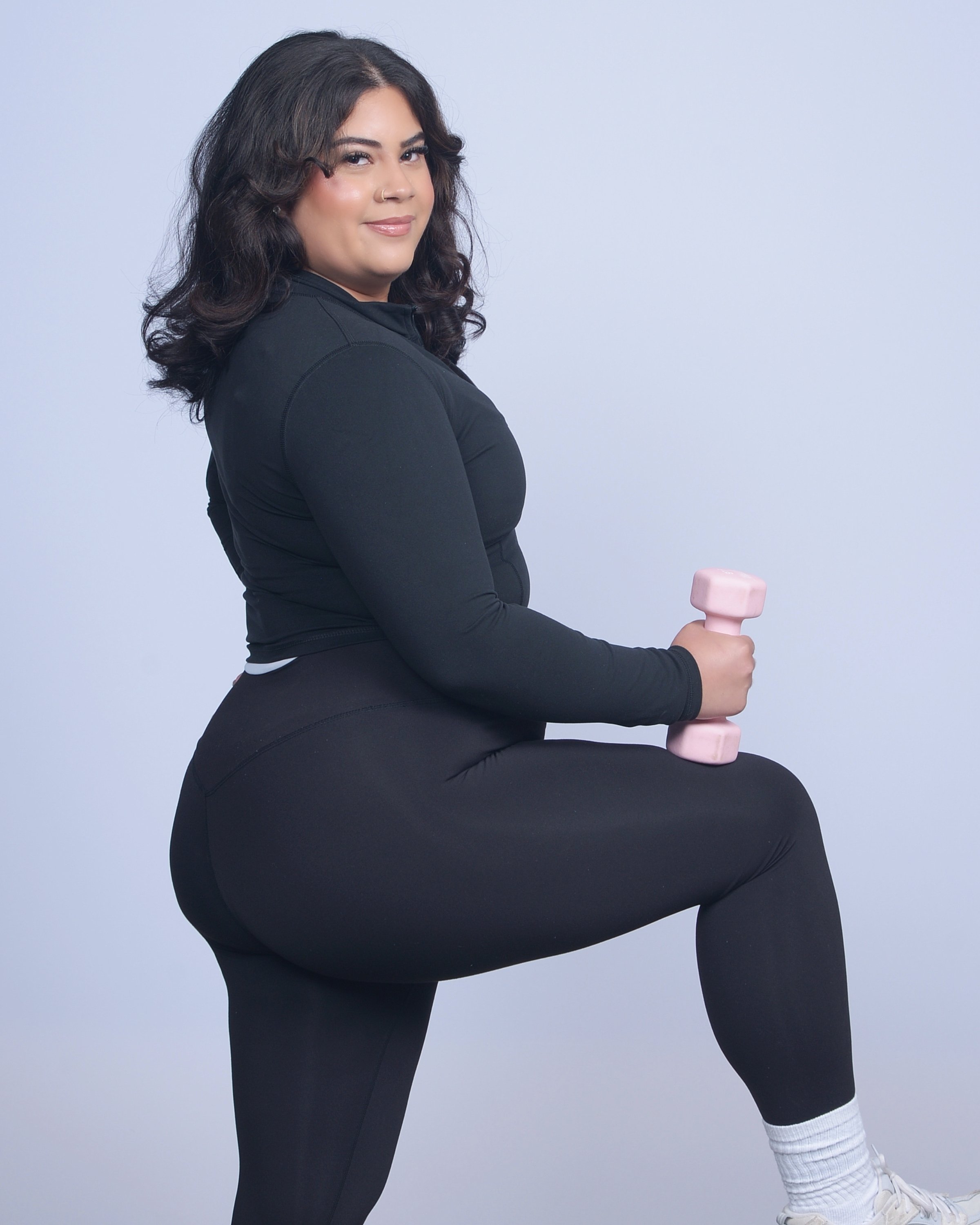 A woman with dark curly hair, wearing black athletic clothing, blonde socks, and white sneakers, sitting on a chair in a squat position holding a pink dumbbell.