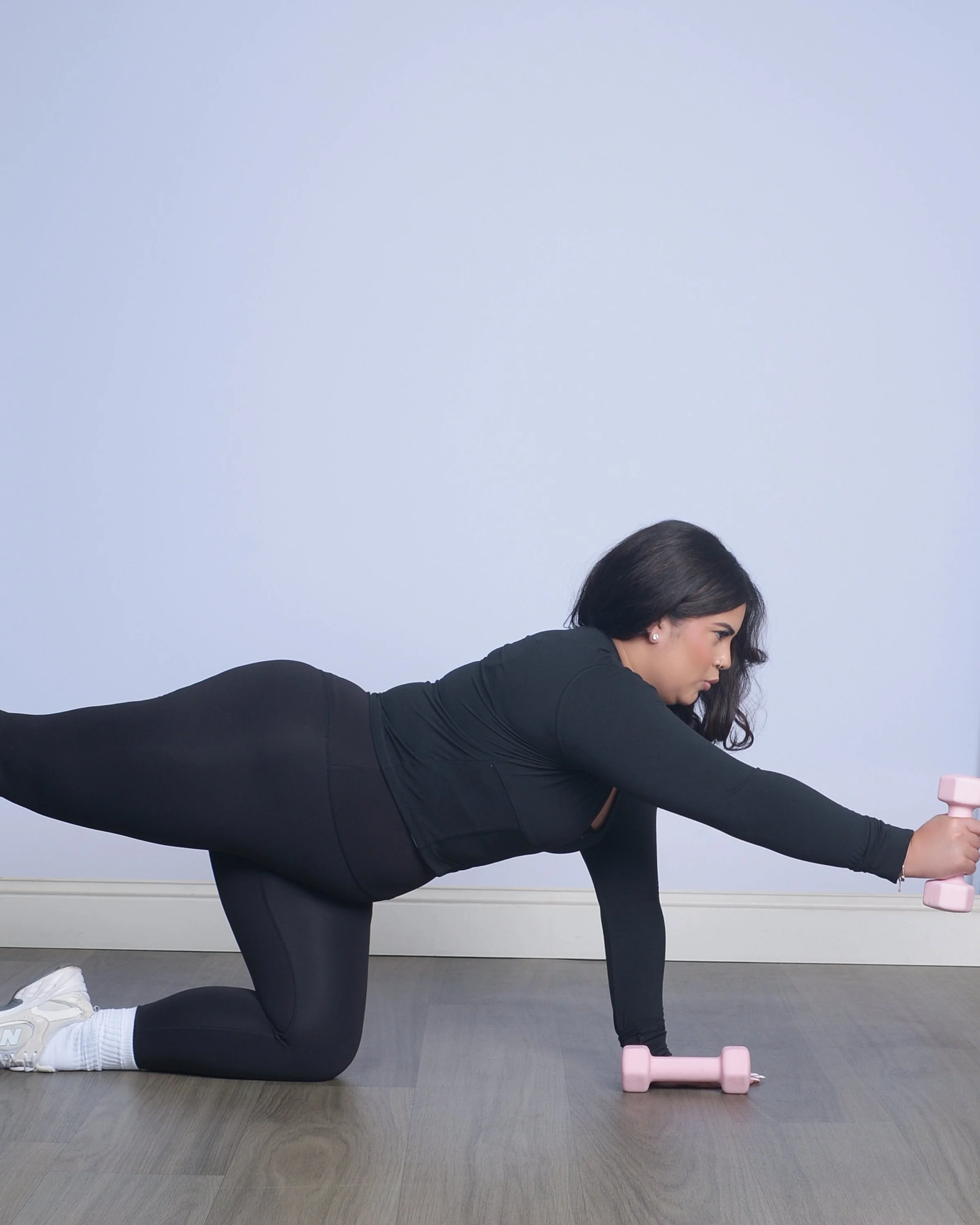 Woman in black athletic wear performing a stretch or yoga pose on her hands and knees in a room with a light blue wall and pink dumbbells.