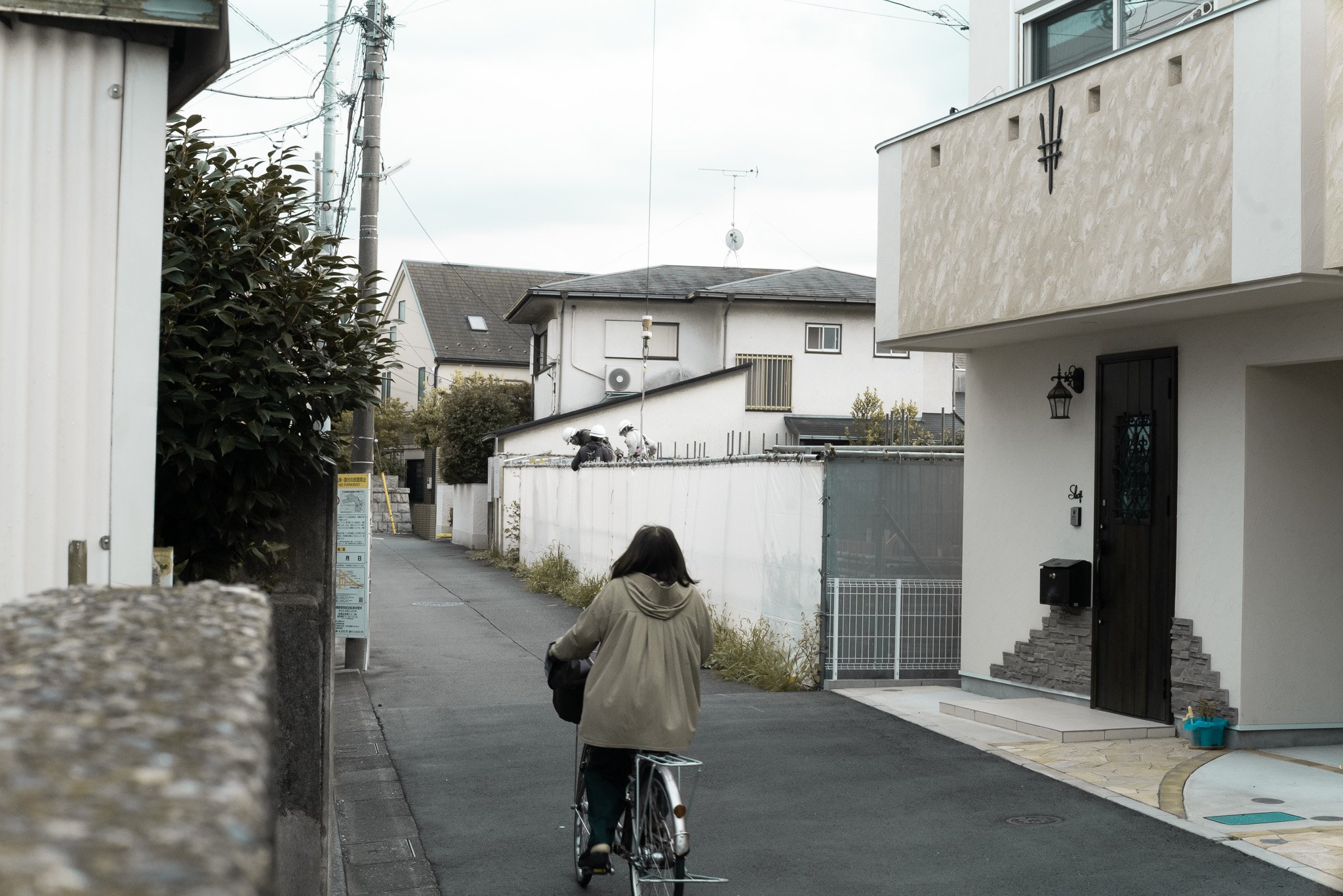 小さな道の左側に木の茂みと電柱があり、右側には白い家と女性が自転車に乗っている様子が映っている。背景には複数の住宅と空が見える。
