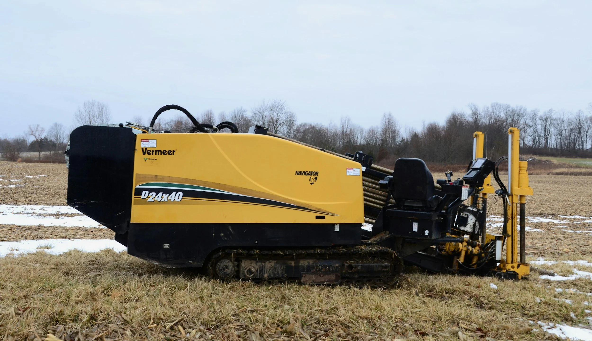 Yellow Vermeer Navigator D24x40 agricultural machine moving through a field with patches of snow, with a cloudy sky and bare trees in the background.