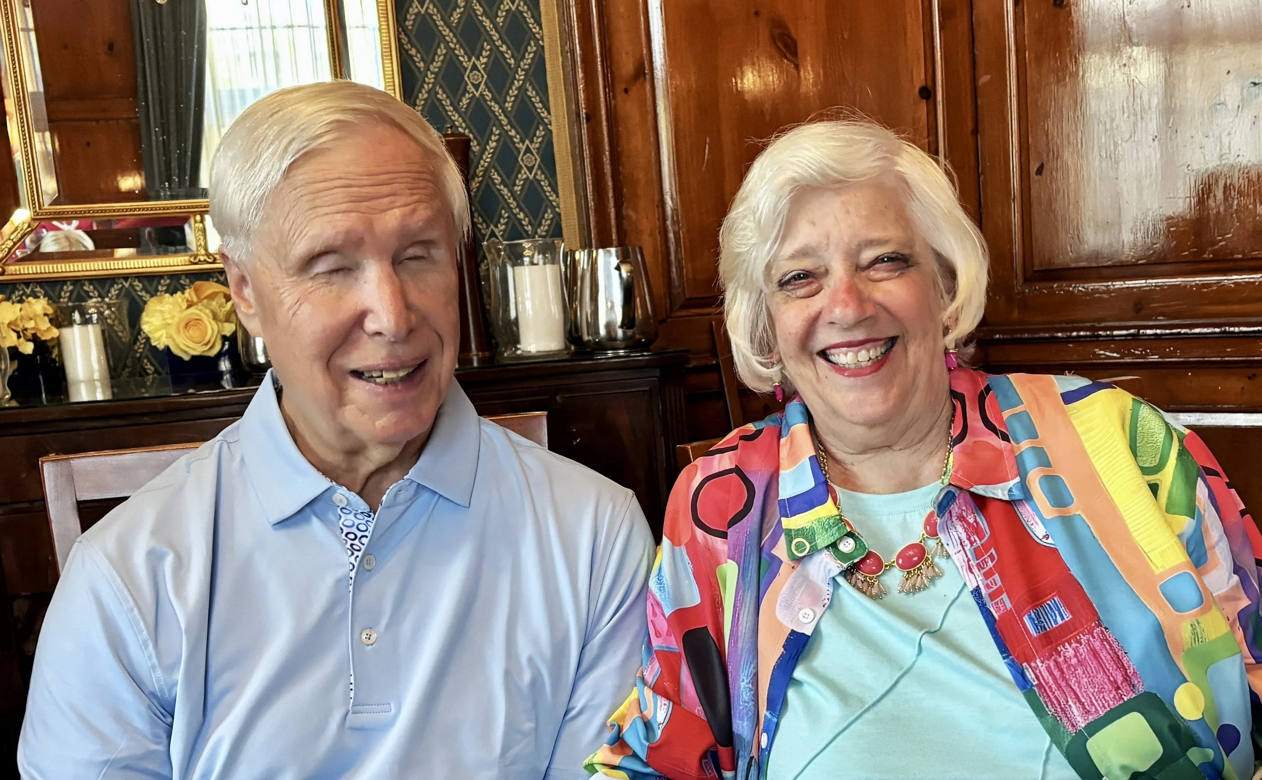 A man and a woman sitting together, both smiling in a room with wooden paneled walls, a mirror, and decorative yellow flowers and candles in the background.
