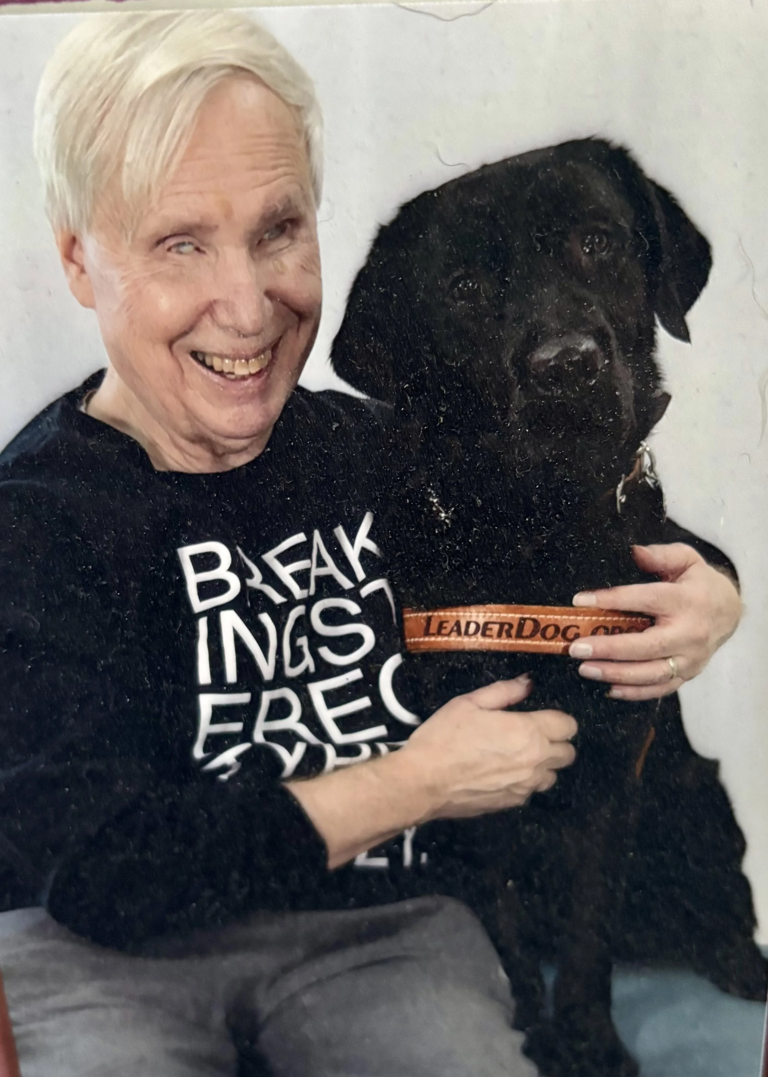 An elderly woman with short white hair smiling and holding a black dog with a collar that reads 'LEADERDOG.' The woman is wearing a black shirt with white text, and the dog is looking towards the camera. The woman appears to be happy, and the background is plain and white.