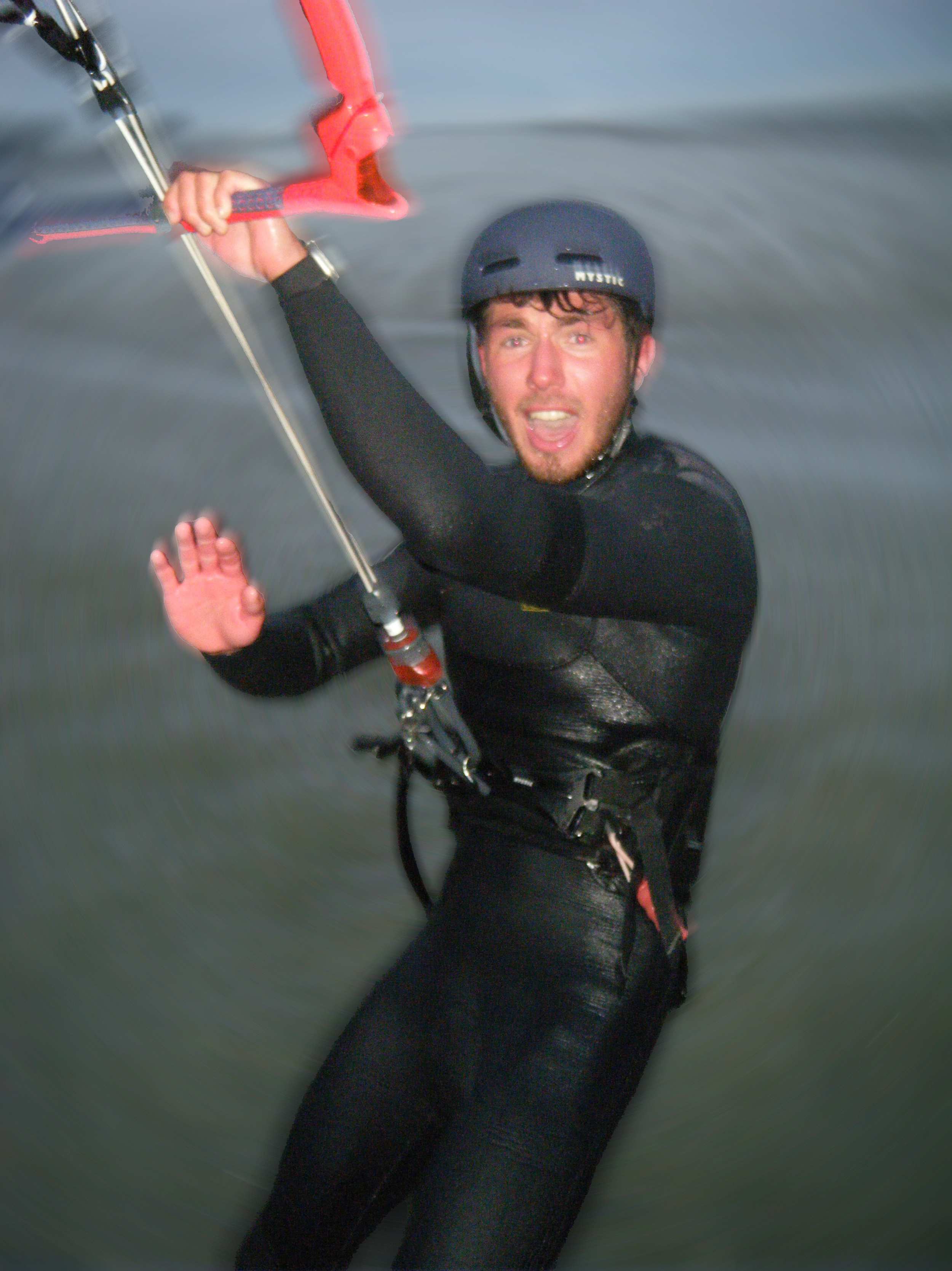 Ein Mann trägt einen Helm und einen Neoprenanzug, hält eine Klampe und befindet sich wahrscheinlich beim Kitesurfen auf Wasser.
