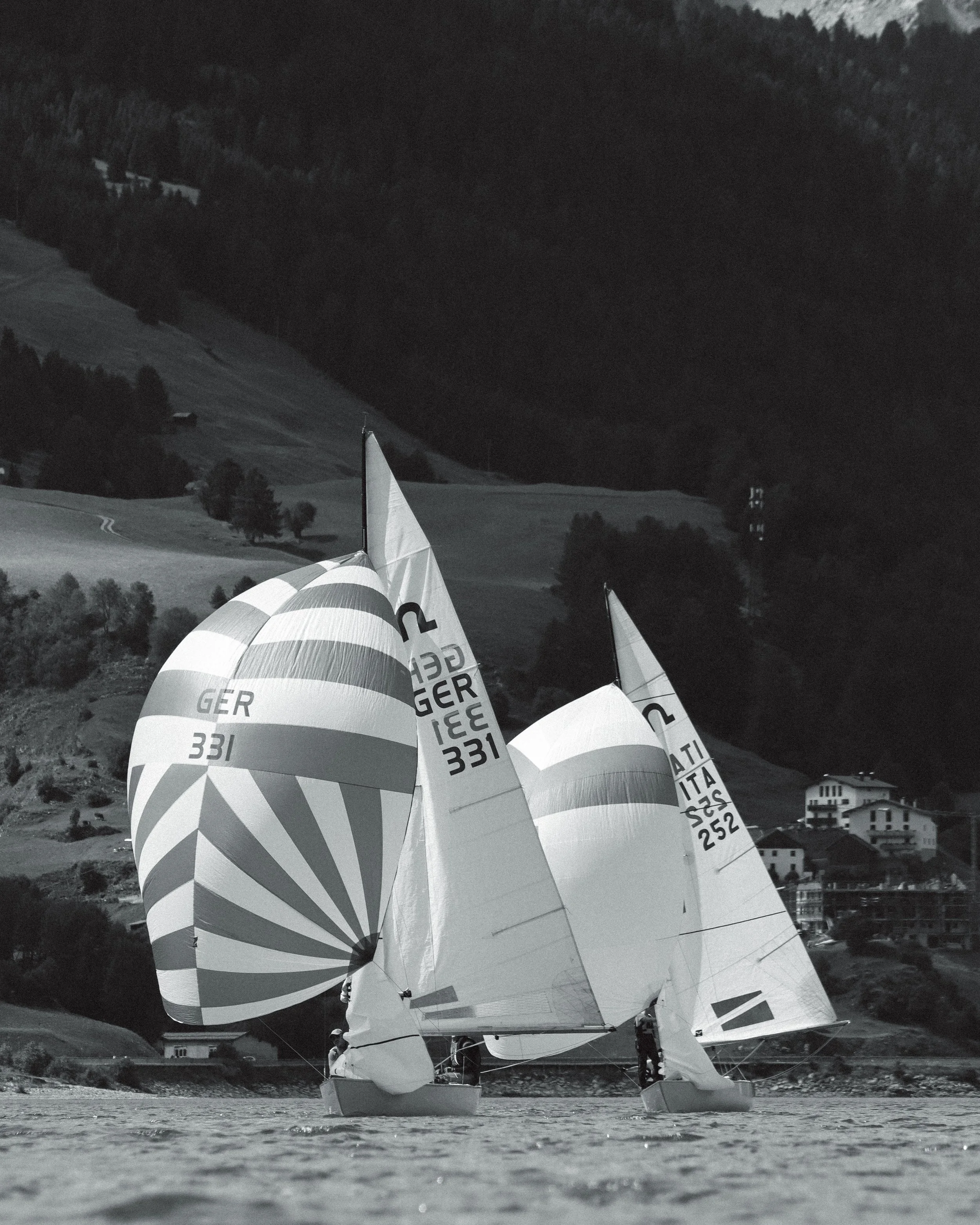 Segelboote mit Windsurfbrettern auf einem Fluss, im Hintergrund Berge und Häuser, schwarz-weißes Bild.