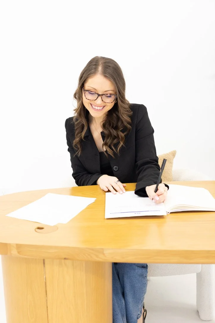 A woman with long wavy brown hair and glasses is smiling while writing in a notebook at a wooden desk. There are papers on the desk, and she is wearing a black blazer and jeans, sitting against a plain white background.