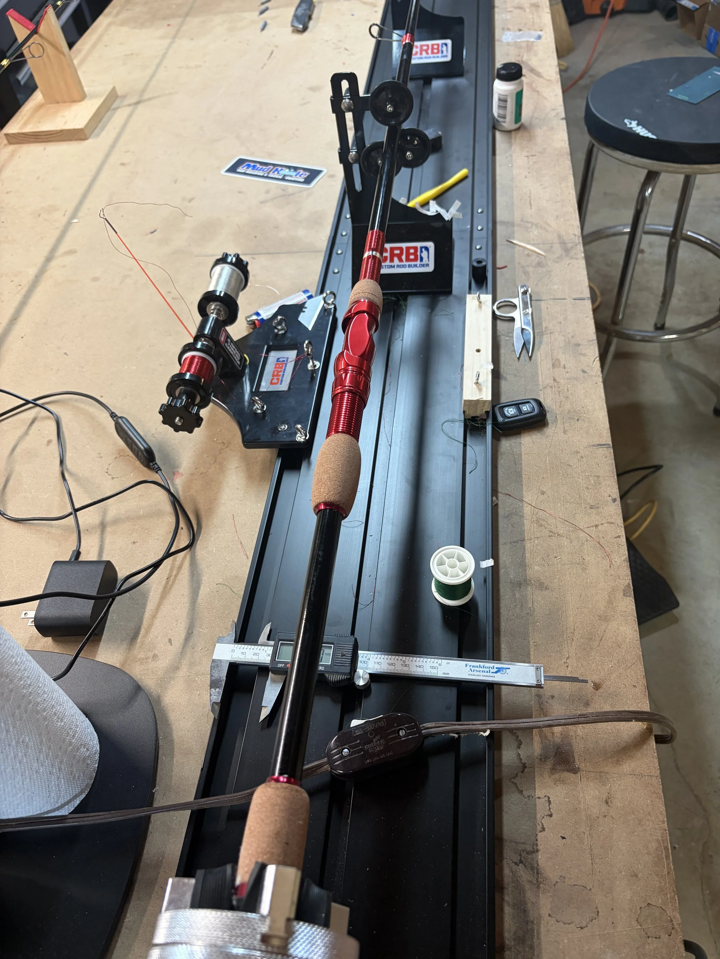 A woodworking workbench with a mounted fishing rod setup, digital scales, scissors, a small shelf, bottle of supplements, and extra fishing reel parts, surrounded by various tools.