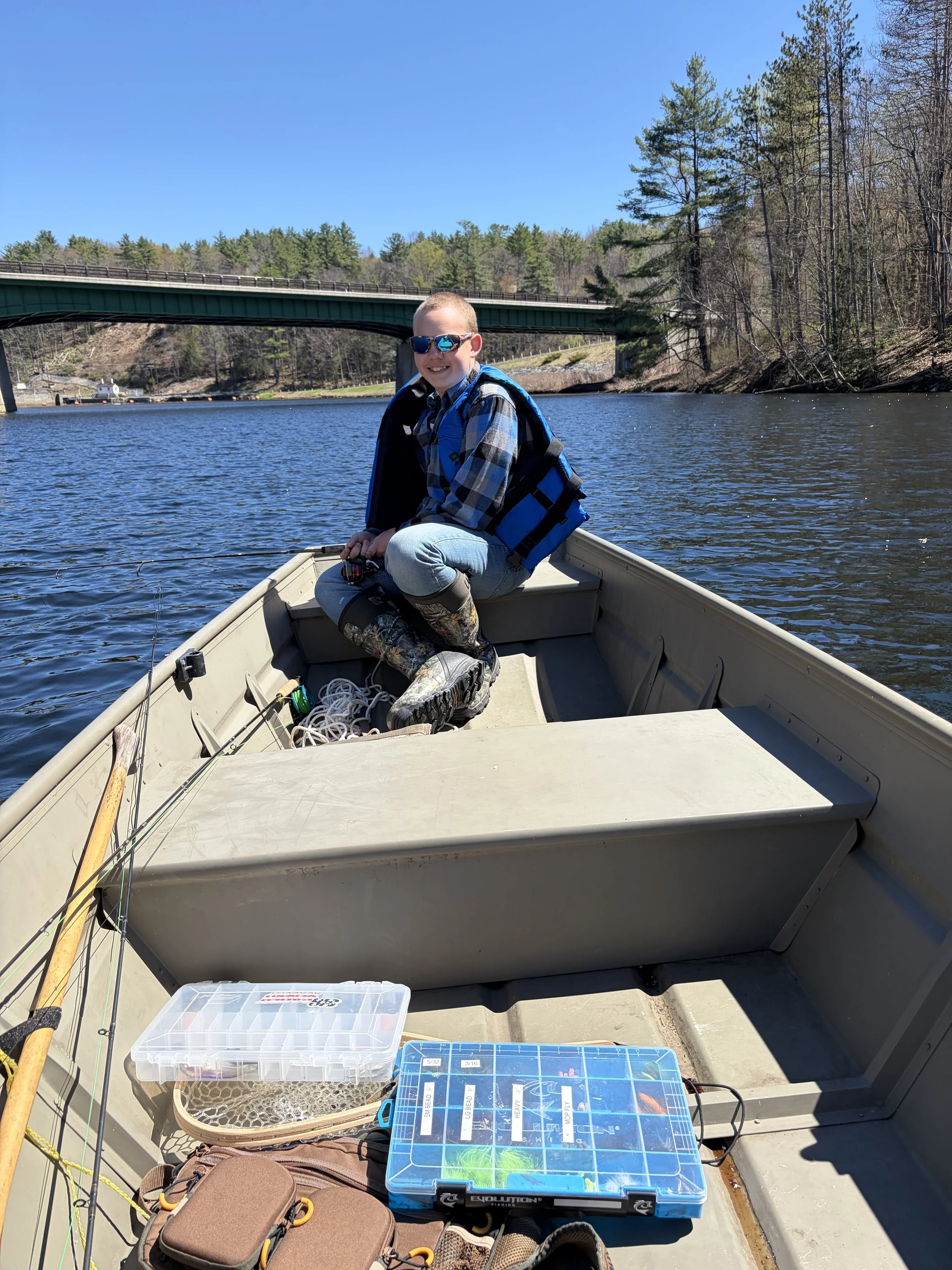 A young boy fishing from a boat on a lake with a bridge and trees in the background, sunny day with clear blue sky.
