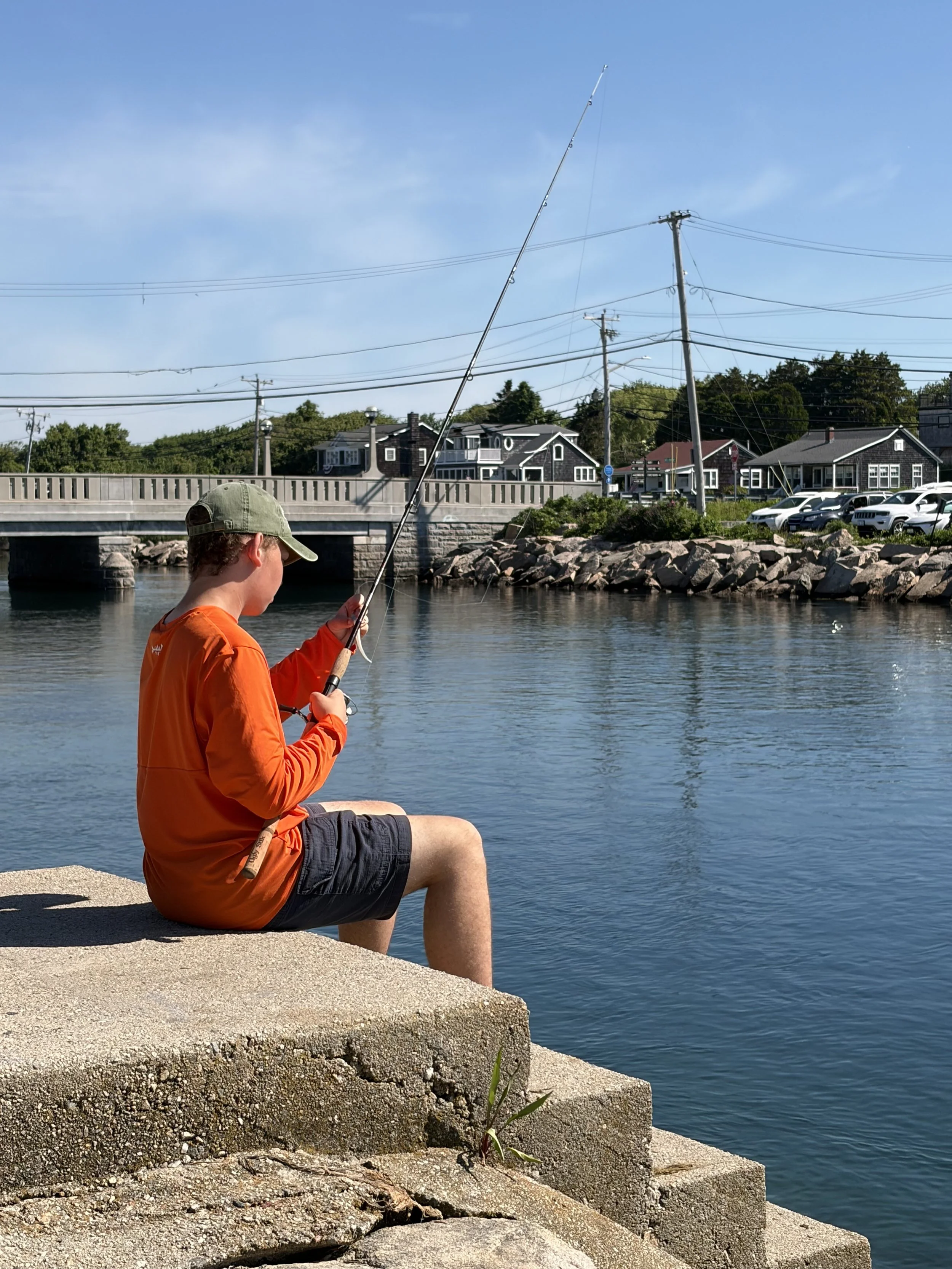 A young man in an orange jacket and dark shorts sitting on a concrete dock at the water's edge, fishing with a pole, with utility poles, houses, and a bridge in the background on a clear day.