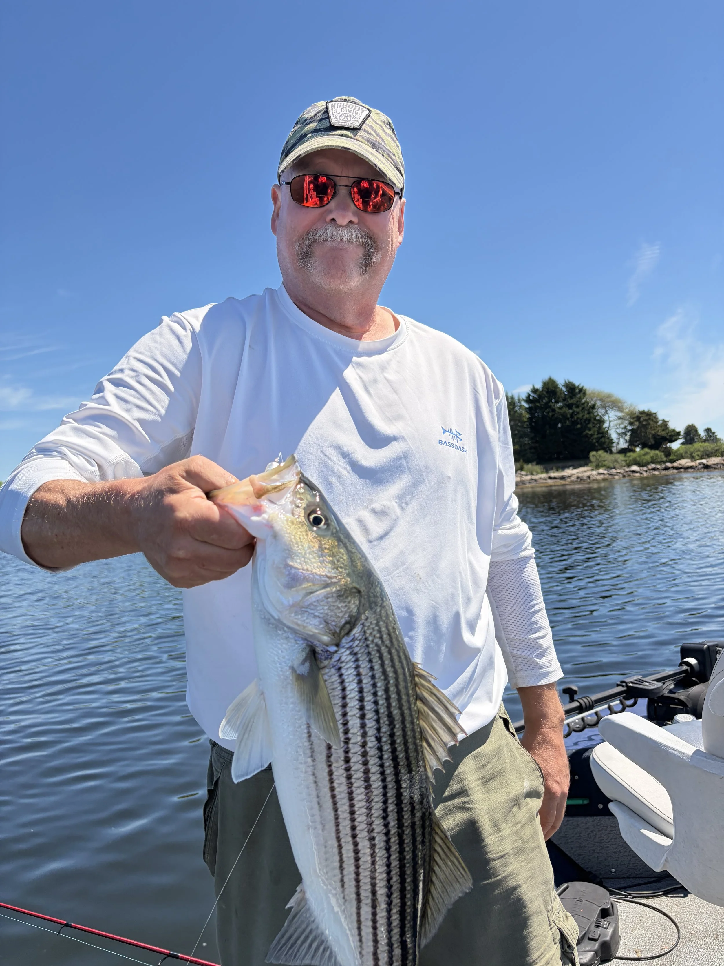 A man holding a large Striped Bass, wearing sunglasses,  on a boat during a sunny day with blue sky and water in the background.