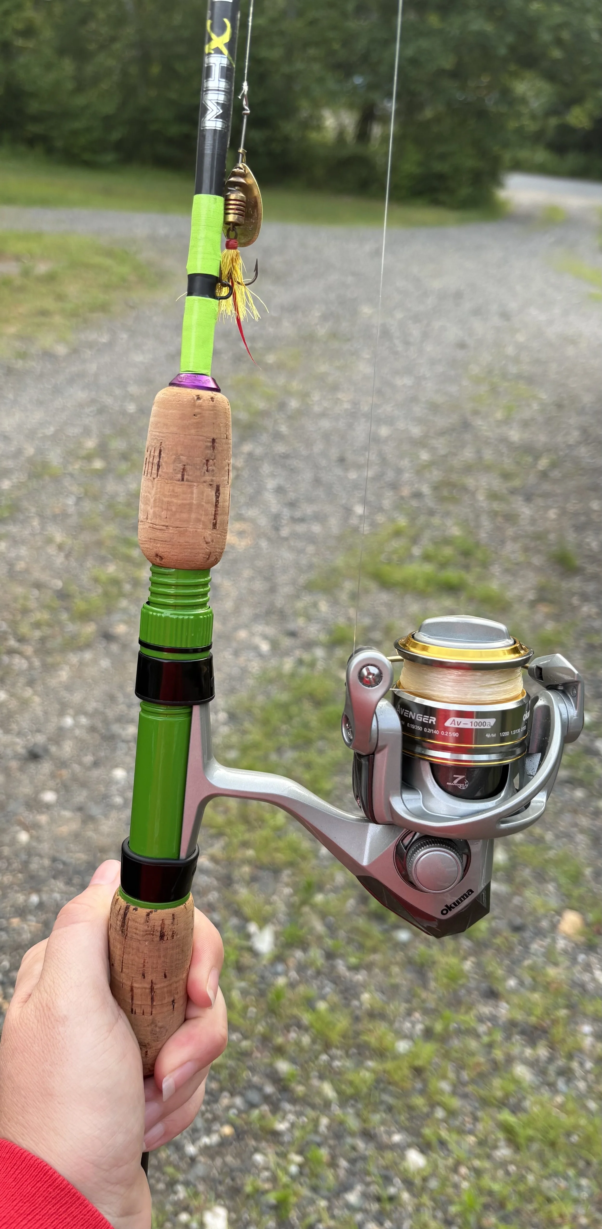 A fishing rod with green accents and a cork handle, attached to a spinning reel, held by a hand near a gravel path with greenery in the background.