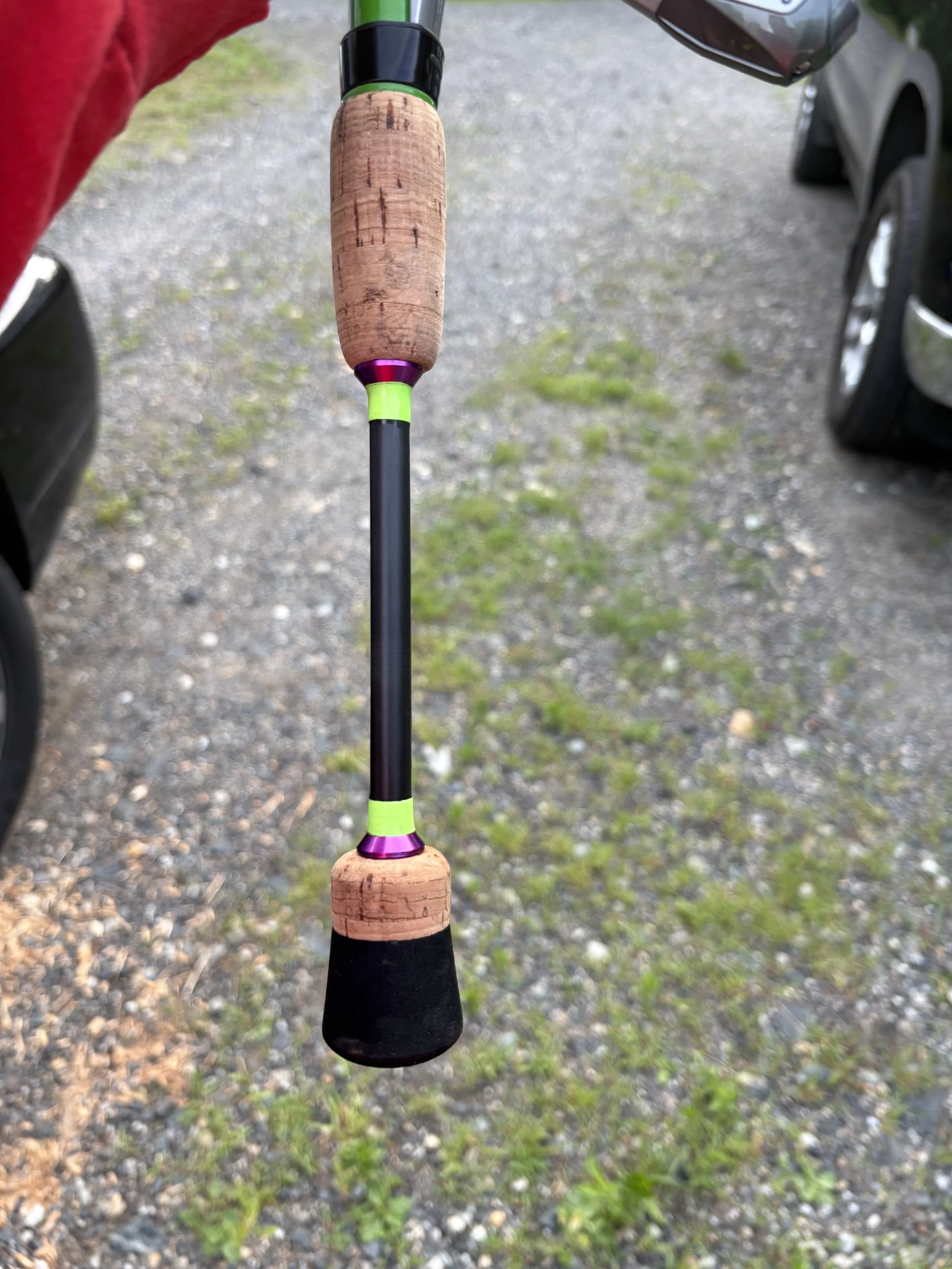 A fishing rod with a cork handle, green and purple accents, and a black reel handle, held by a person wearing a red shirt, against a gravel ground with grass patches and vehicles nearby.