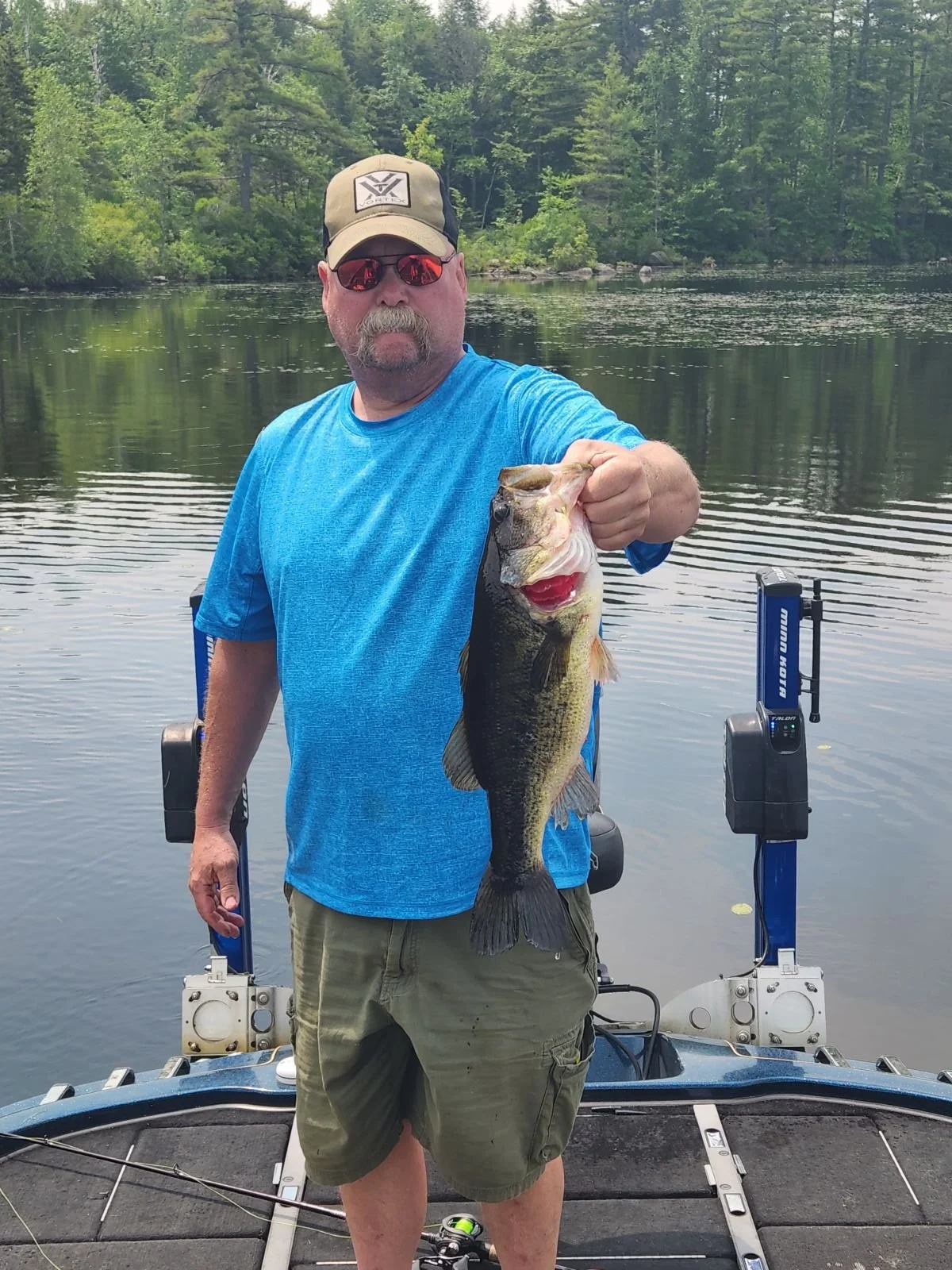A man standing on a boat holding a large bass fish he caught, with a lake and trees in the background.