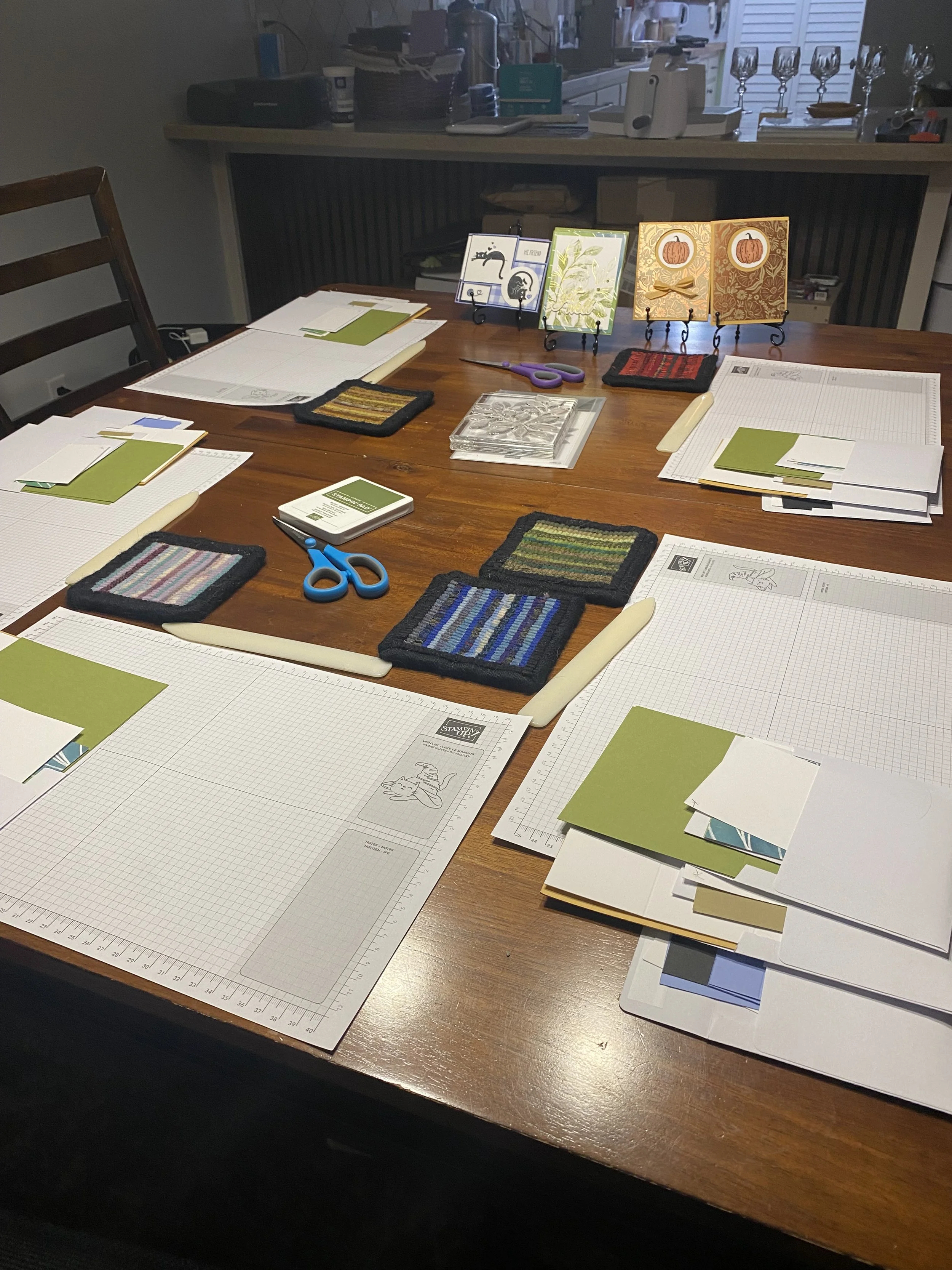 A wooden table set up for crafting, with paper, scissors, color samples, and decorative greeting cards displayed on small stands. Various crafting supplies are arranged around the table, with a kitchen in the background.