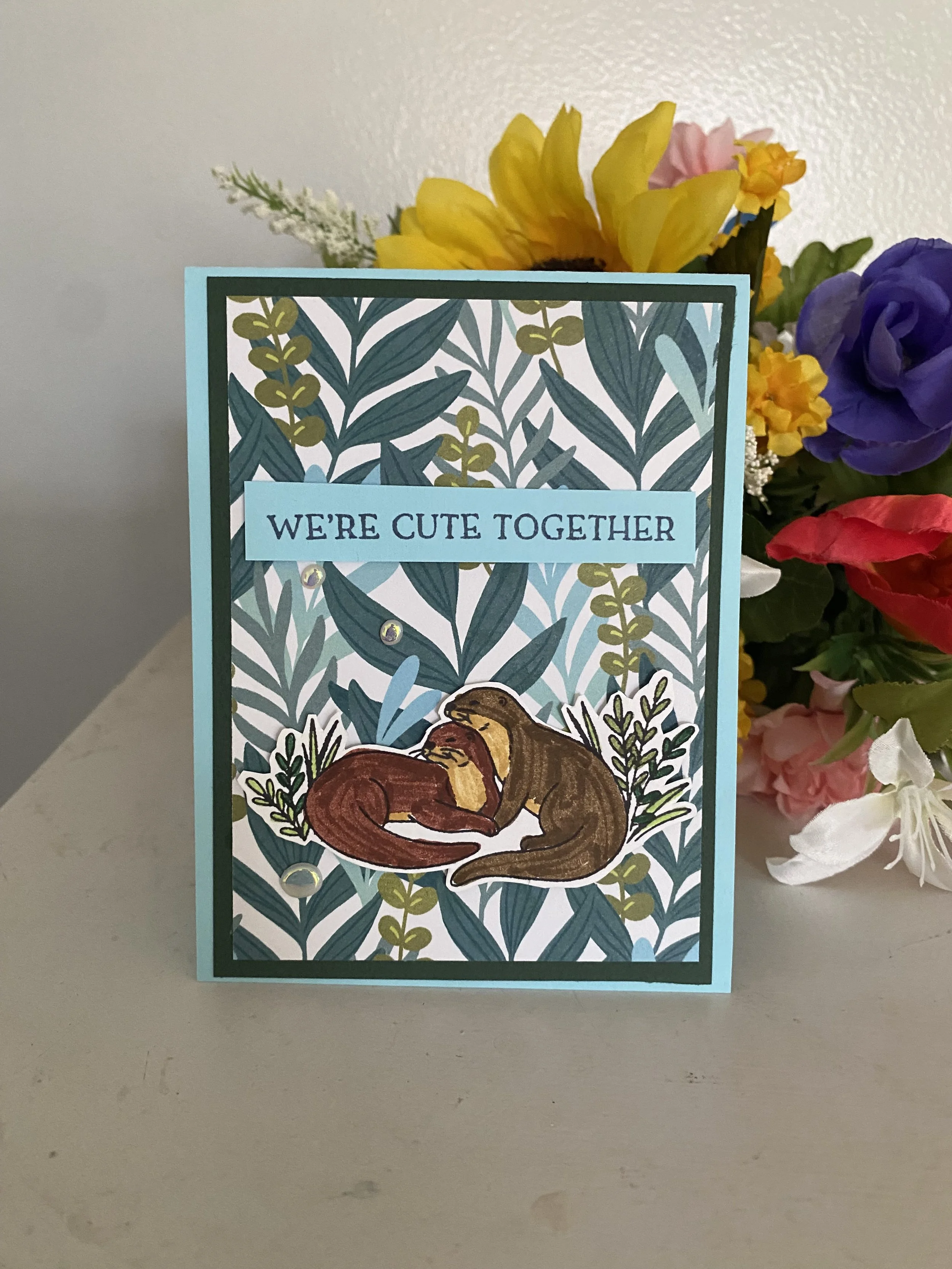 A colorful greeting card with the phrase "We're cute together" and a bear hugging a fox, set against a leafy background, with a bouquet of flowers in the background.