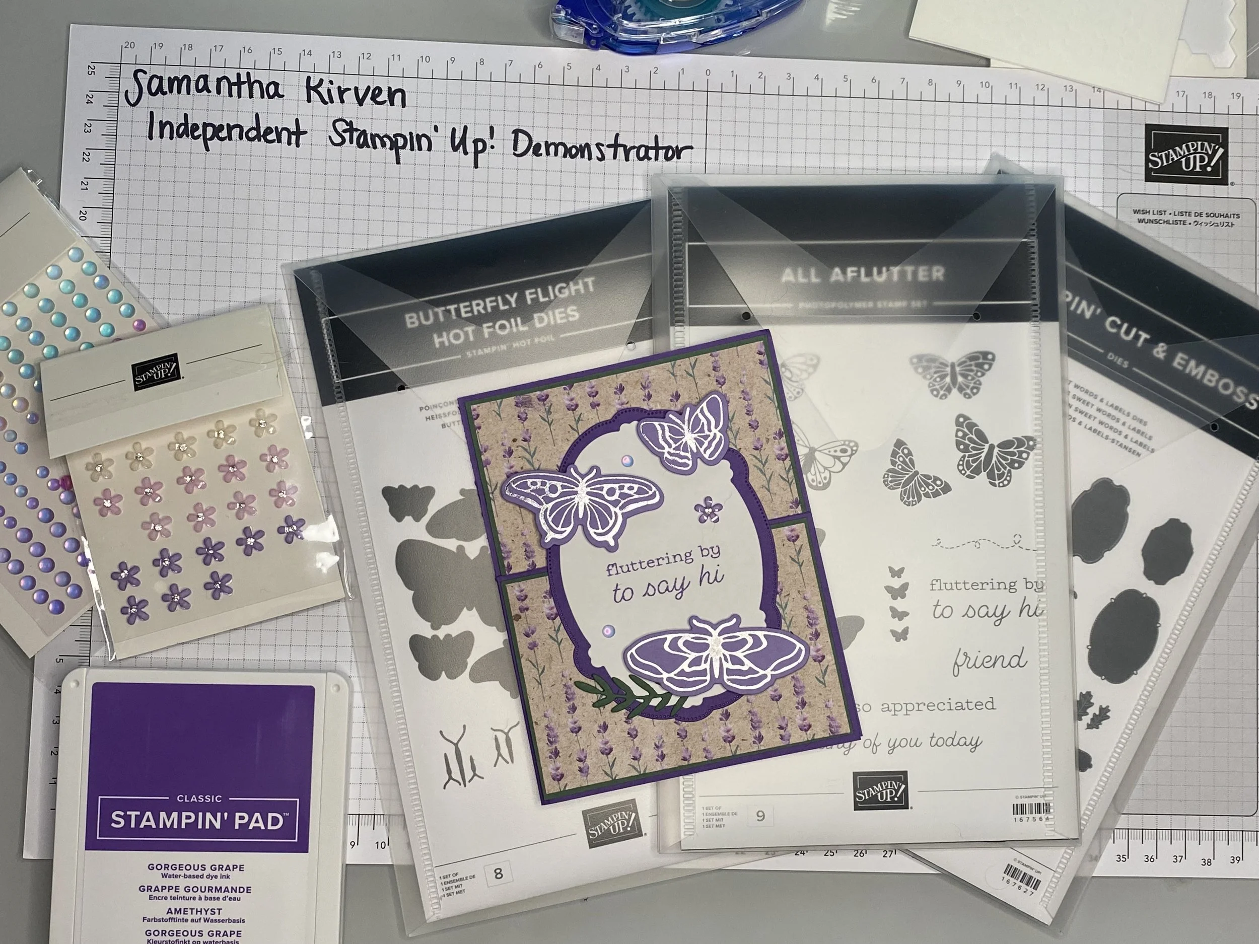 Crafting supplies including sticker sheets with butterflies and flowers, a purple ink pad labeled 'Gorgeous Grape,' stamping blocks, and a paper organizer on a grid mat.