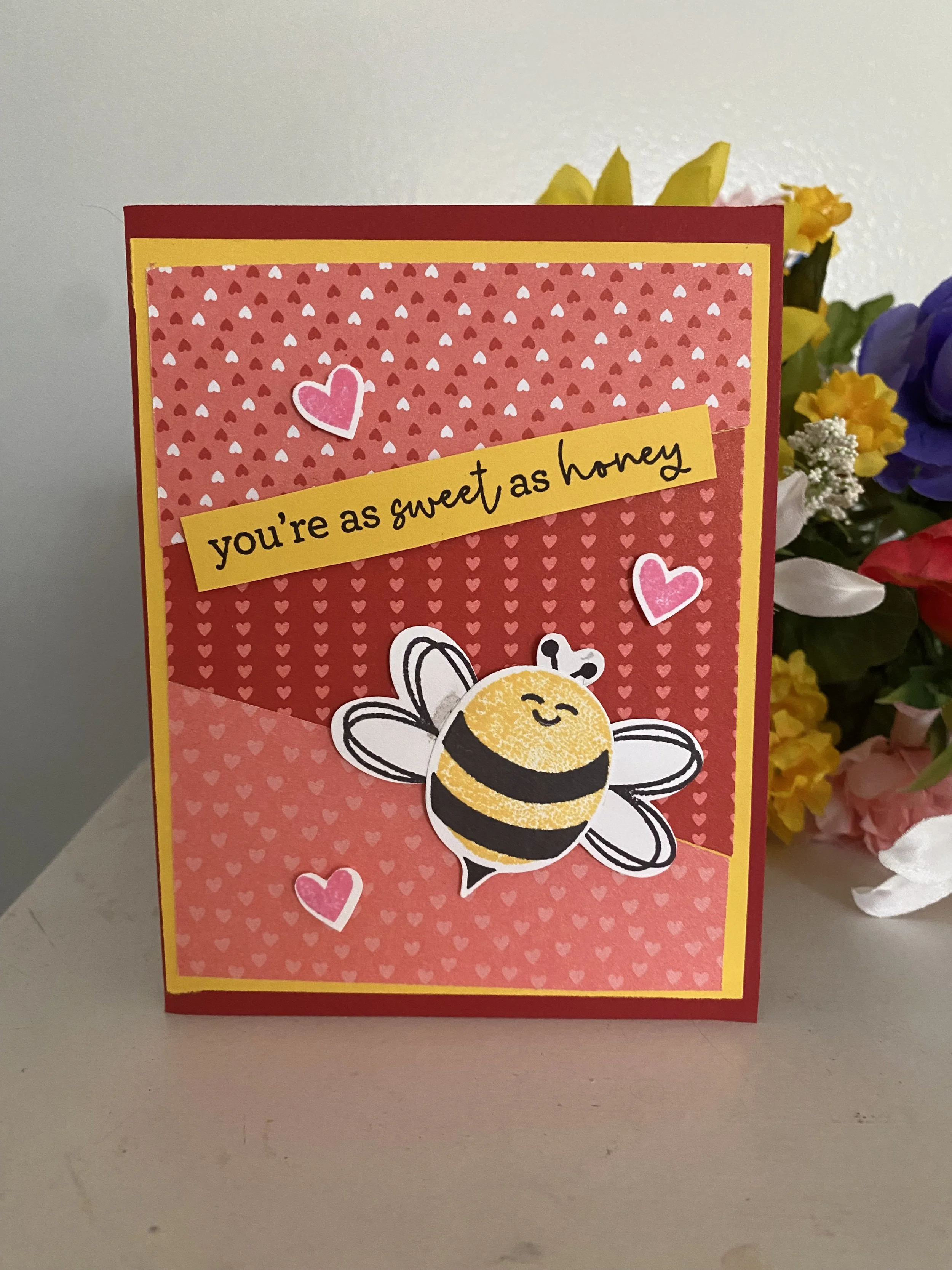 A colorful Valentine's Day card with a honeybee illustration, pink and red hearts, and the phrase 'you're as sweet as honey' on a yellow banner, placed on a grey surface next to a bouquet of mixed flowers.