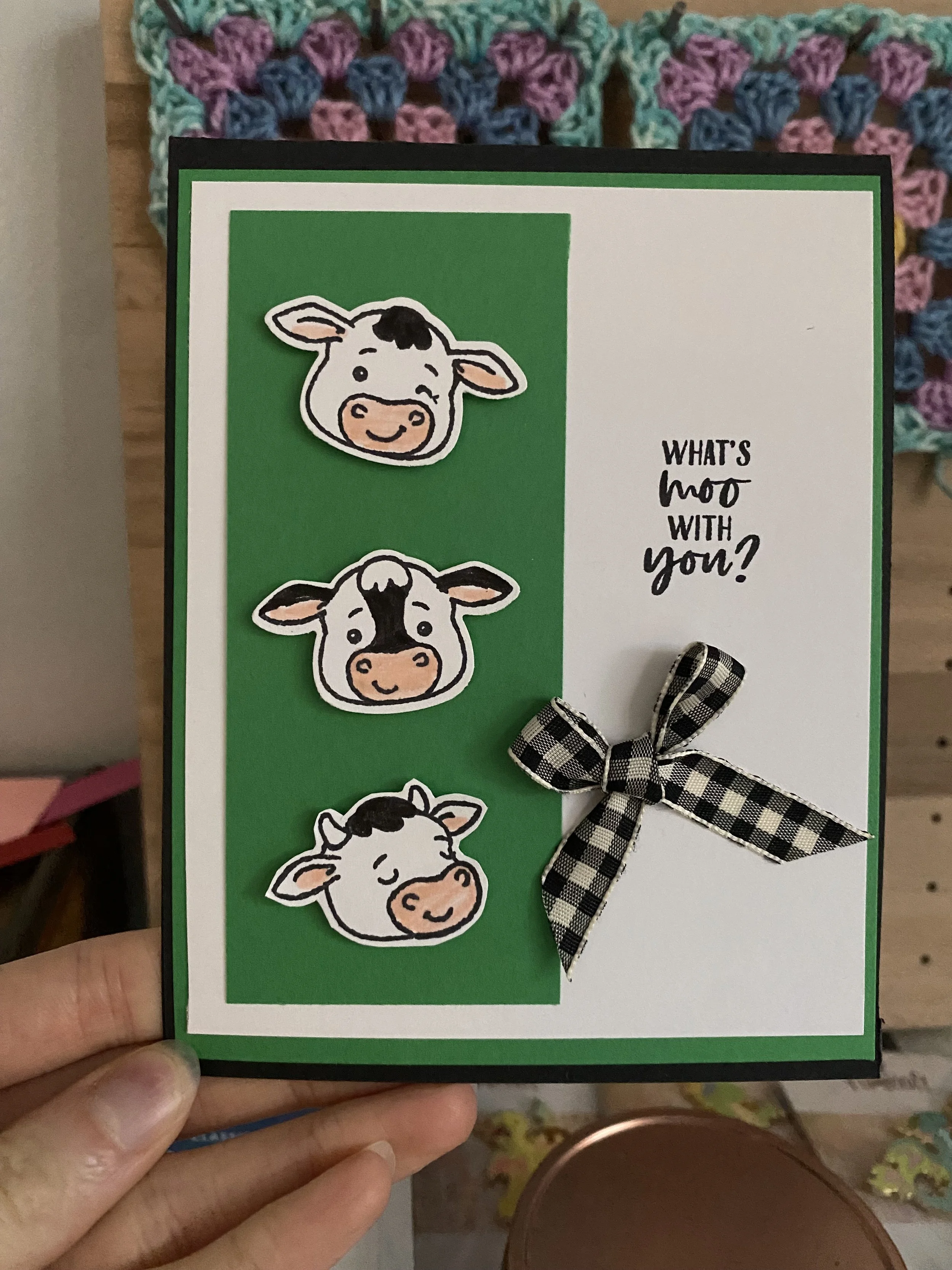 A person holding a small card with cartoon cow faces and a gingham ribbon bow. The card has a green and white background with the text 'WHAT'S moo WITH you?' on it.