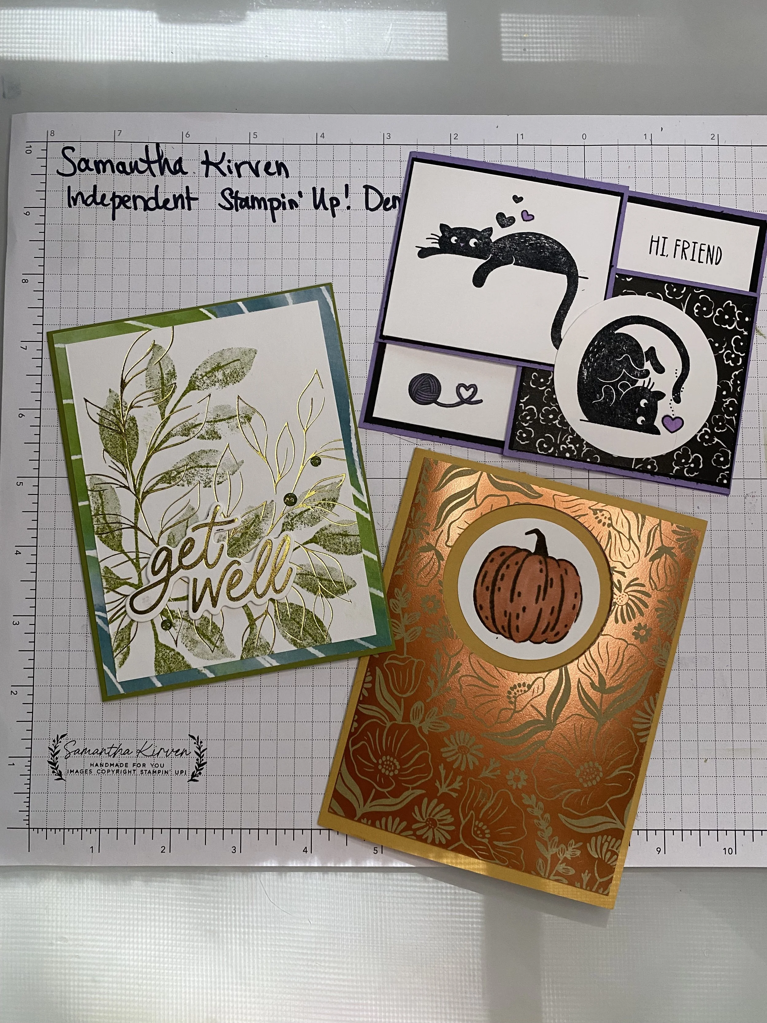Several handmade greeting cards on a grid-lined surface. One card has a floral pattern with the words 'get well' in gold lettering. Another card features a black cat with hearts above it, and the text 'HL FRIEND'. A third card depicts a pumpkin with a floral background.