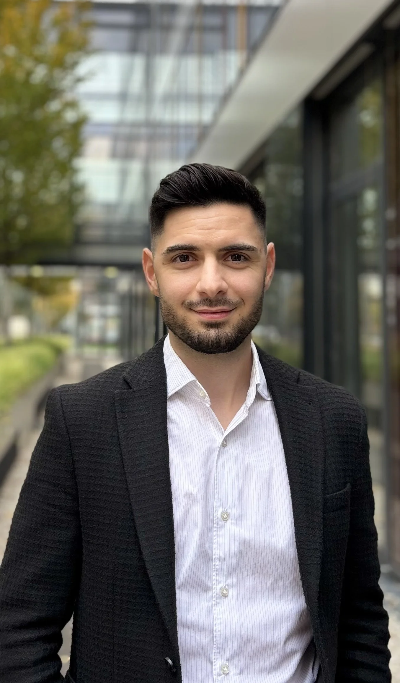 Ein professionell aussehender Mann in Businesskleidung steht vor einem modernen Bürogebäude aus Glas, umgeben von Bäumen, im Freien.