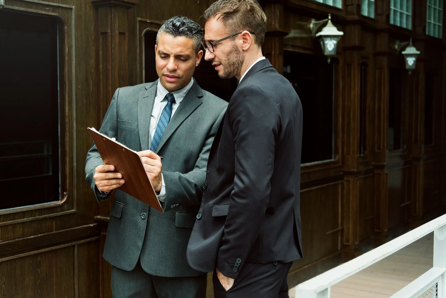 Two men in business suits standing and looking at a clipboard together in an ornate wood-paneled room.