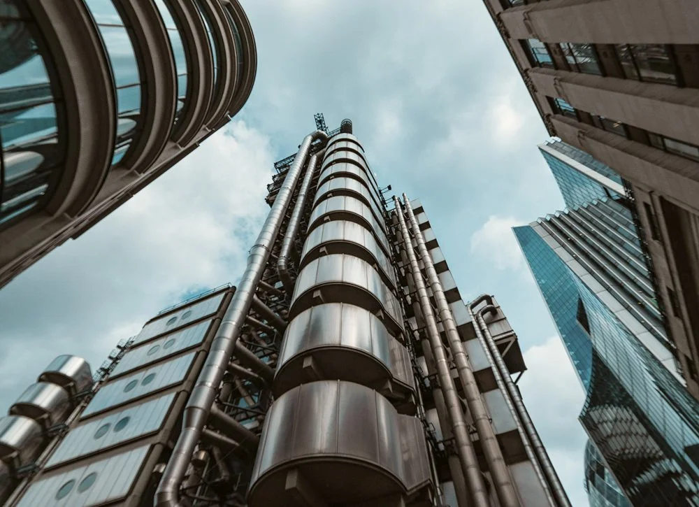 Looking up at a collection of modern high-rise buildings with glass and metal facades, including a prominent building with cylindrical metallic structures and various pipes and frameworks, under a partially cloudy sky.