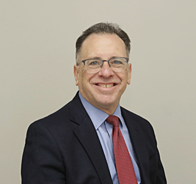 A smiling man in a suit and tie, wearing glasses, against a plain beige wall.