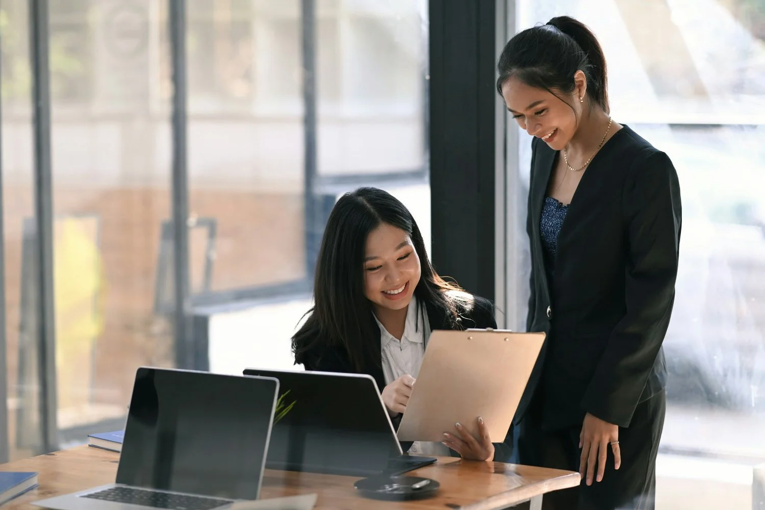 Two women in business attire look at a tablet and smile in a modern office with large windows and laptops on the table.