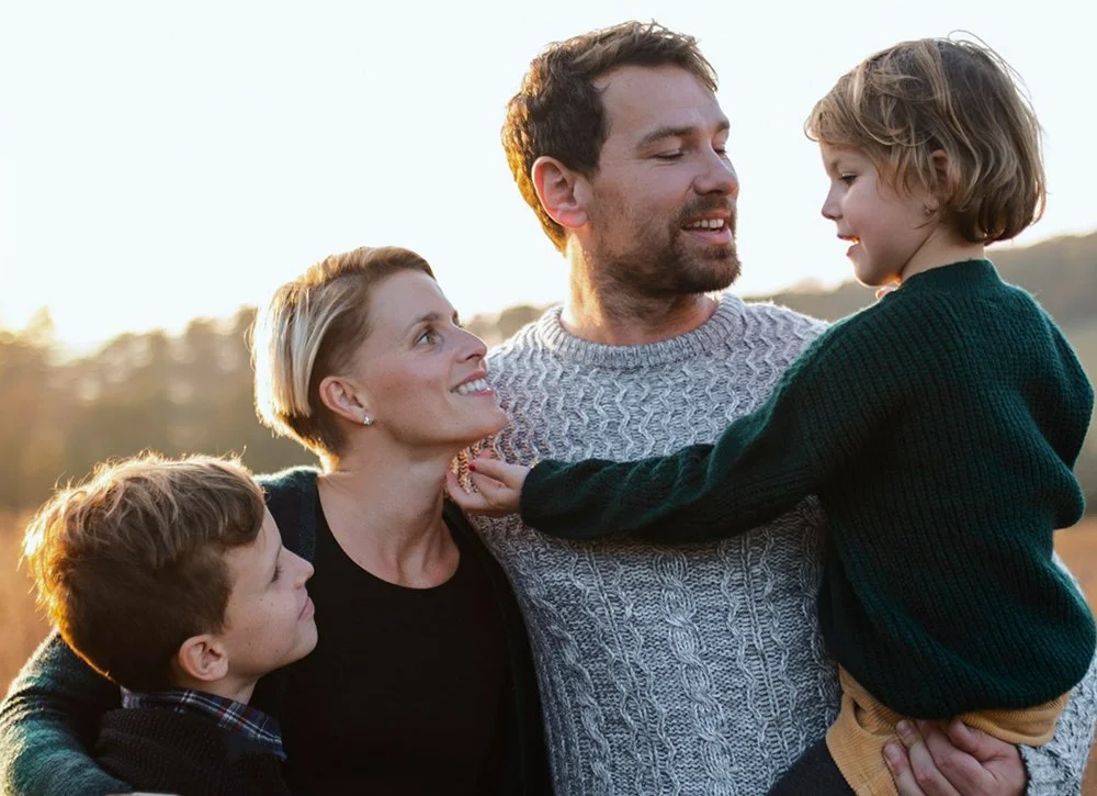 A family of four outdoors during sunset, smiling and interacting; father holding a young boy, mother smiling at a girl who is reaching out to touch her face.