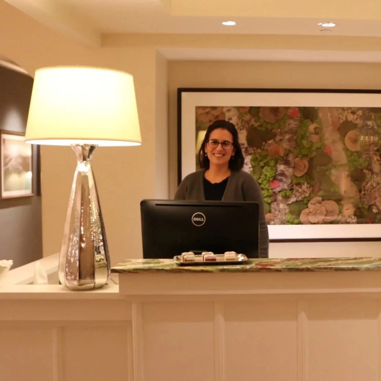 Smiling woman with glasses behind a reception desk with a laptop, a table lamp, and a framed artwork on the wall.